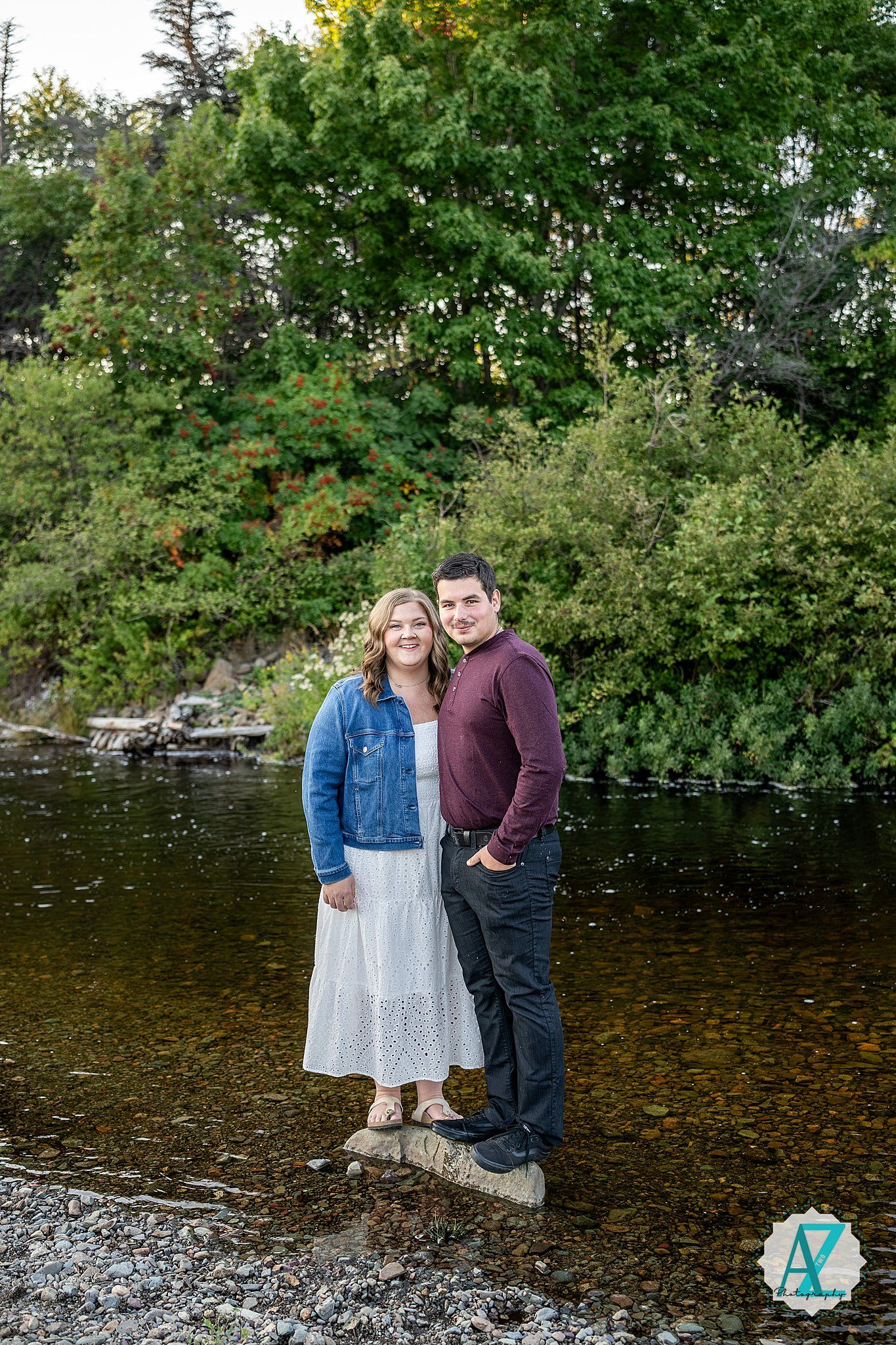 Couple standing together by a serene lake with autumn trees in the background, displaying natural outdoor setting.