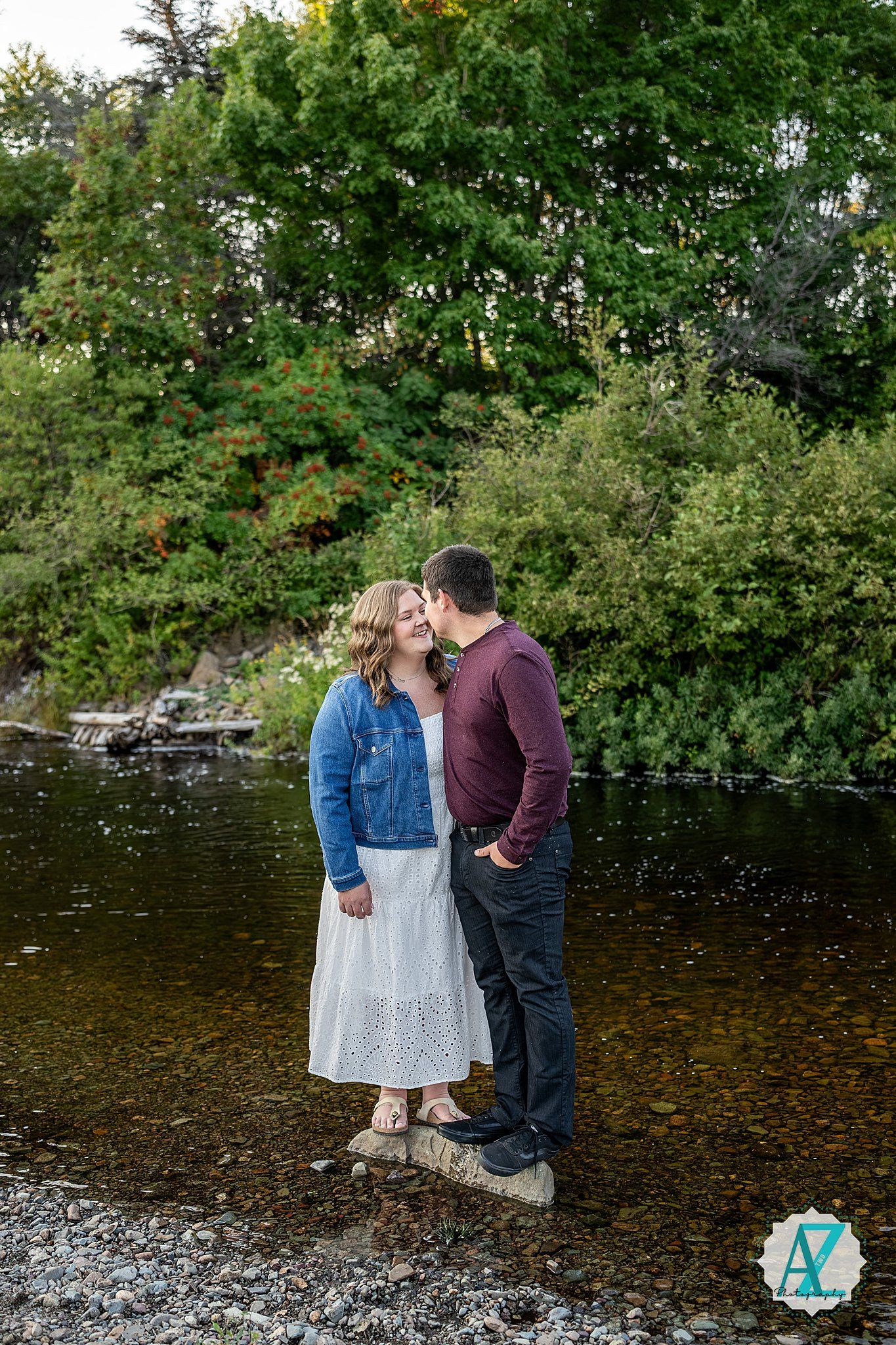 A couple shares a romantic moment standing on a rocky riverbank surrounded by green trees and autumn foliage.
