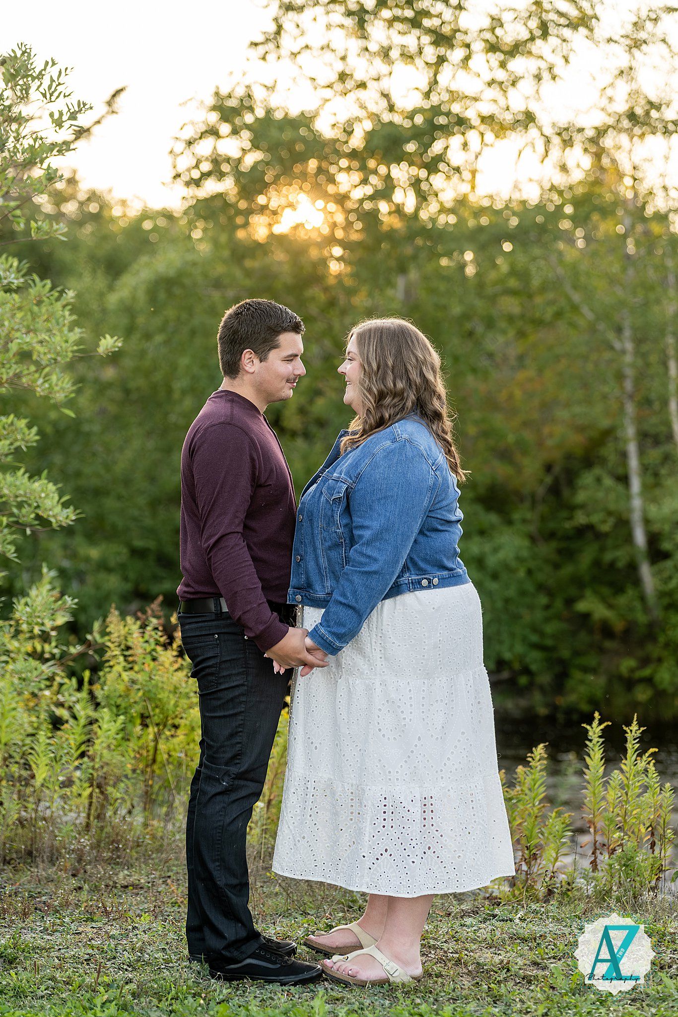 A couple sits together in a meadow at sunset wearing denim and dark clothing with white dress details.