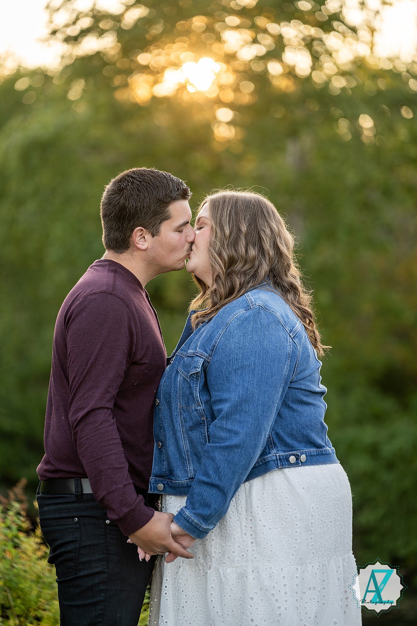 A couple shares a romantic kiss at sunset while standing together in a garden setting against golden light.