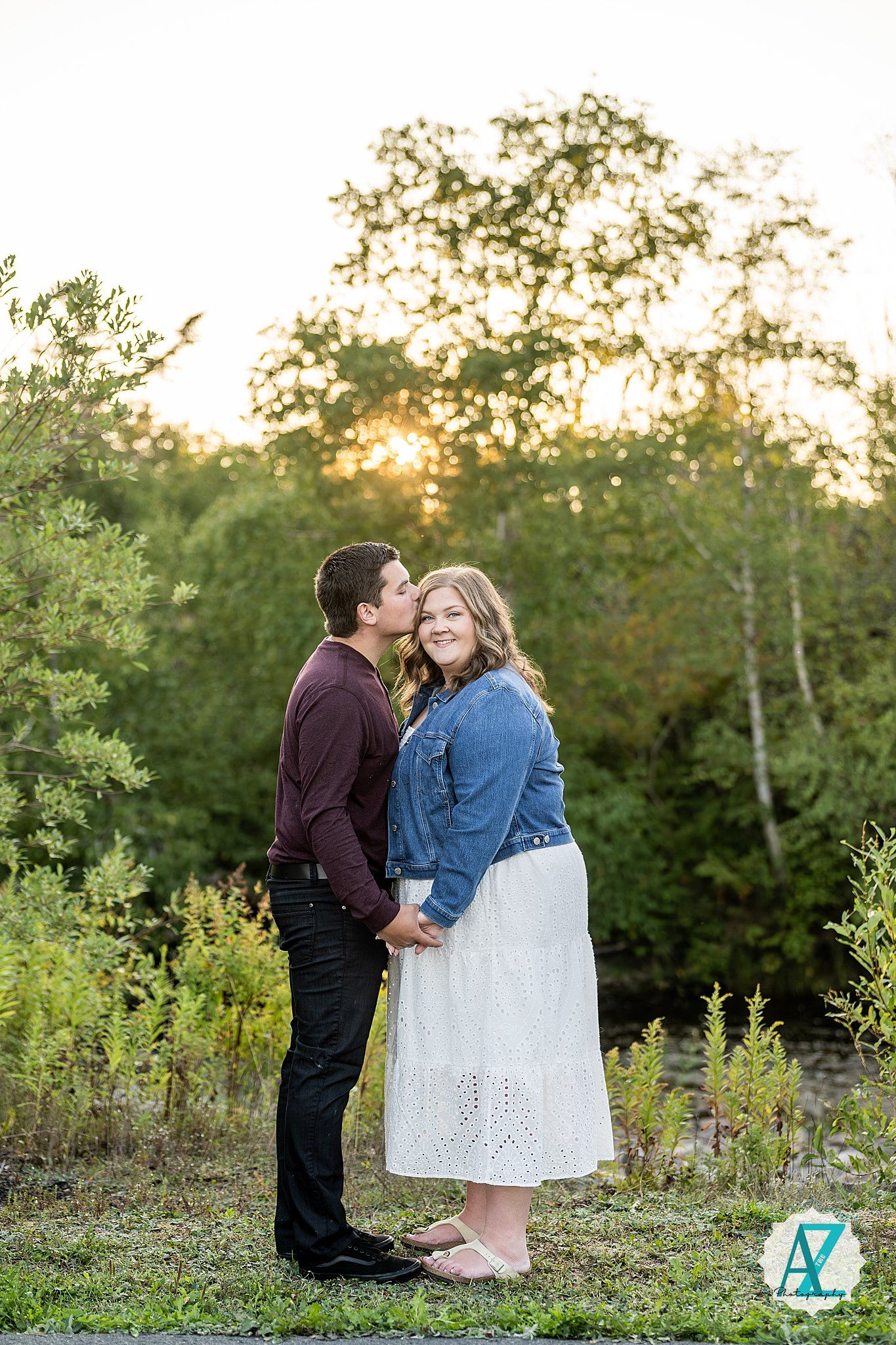 A romantic couple shares a tender moment at sunset in a natural outdoor setting with trees and greenery.