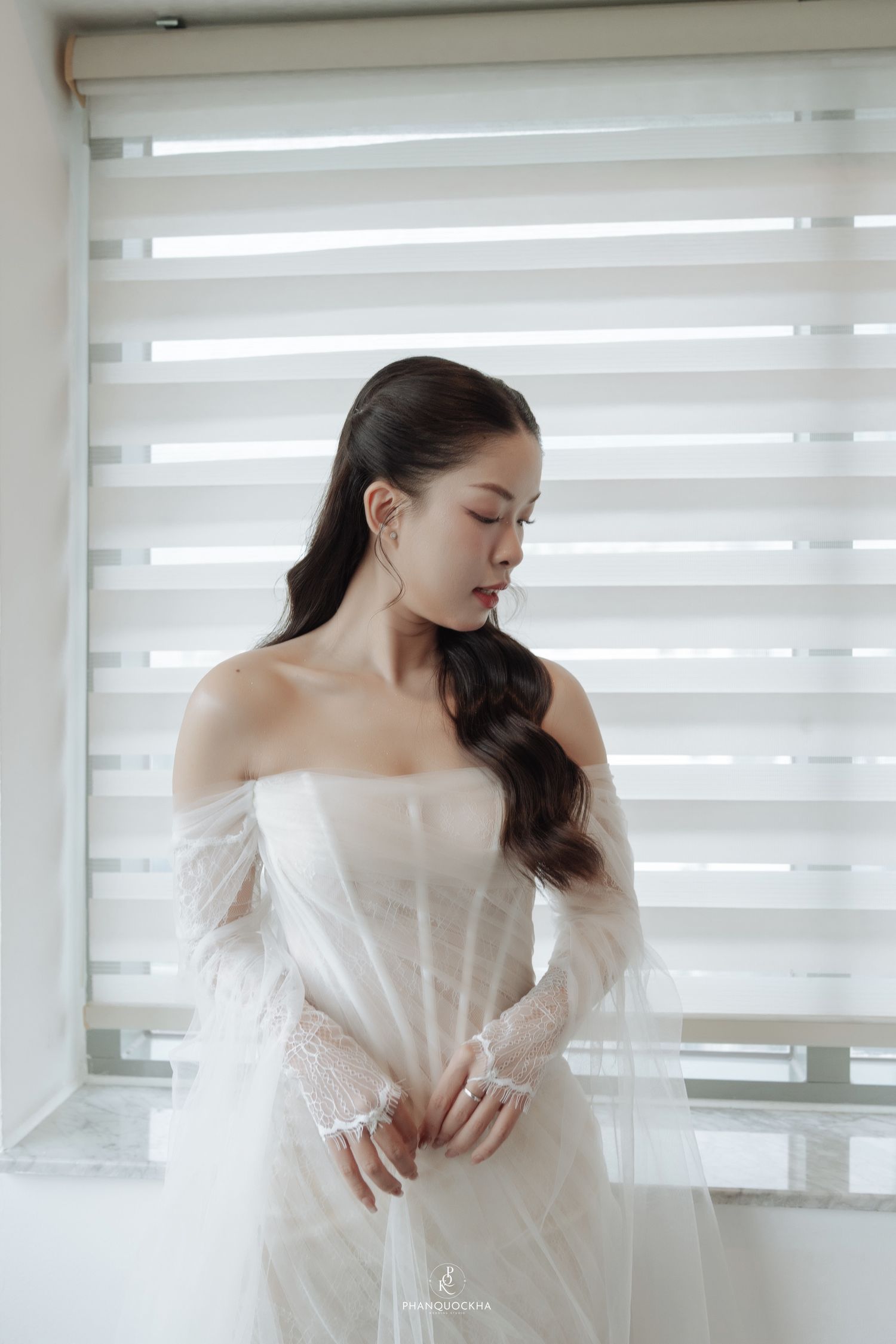 Asian bride in off-shoulder white lace wedding dress looking down by a striped window blind.