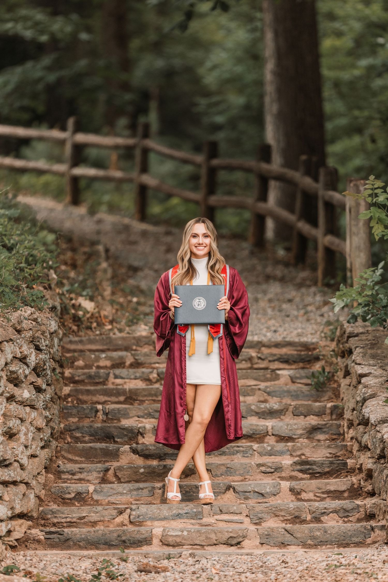 Person in burgundy jacket holding laptop poses on stone stairs in wooded outdoor setting.
