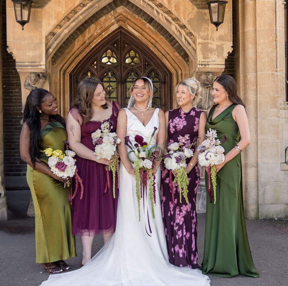 Bride with four bridesmaids in green and purple gowns holding bouquets outside a stone church.
