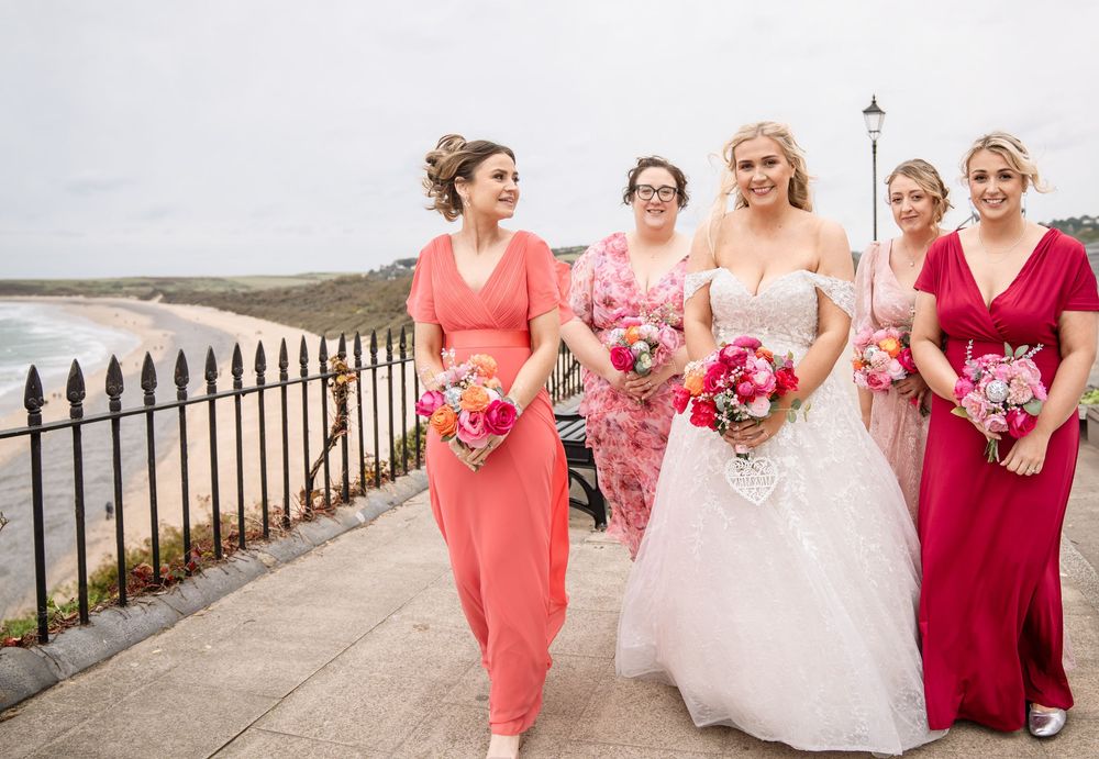 Wedding party in coral and red dresses walking along coastal pathway with bouquets of pink flowers.