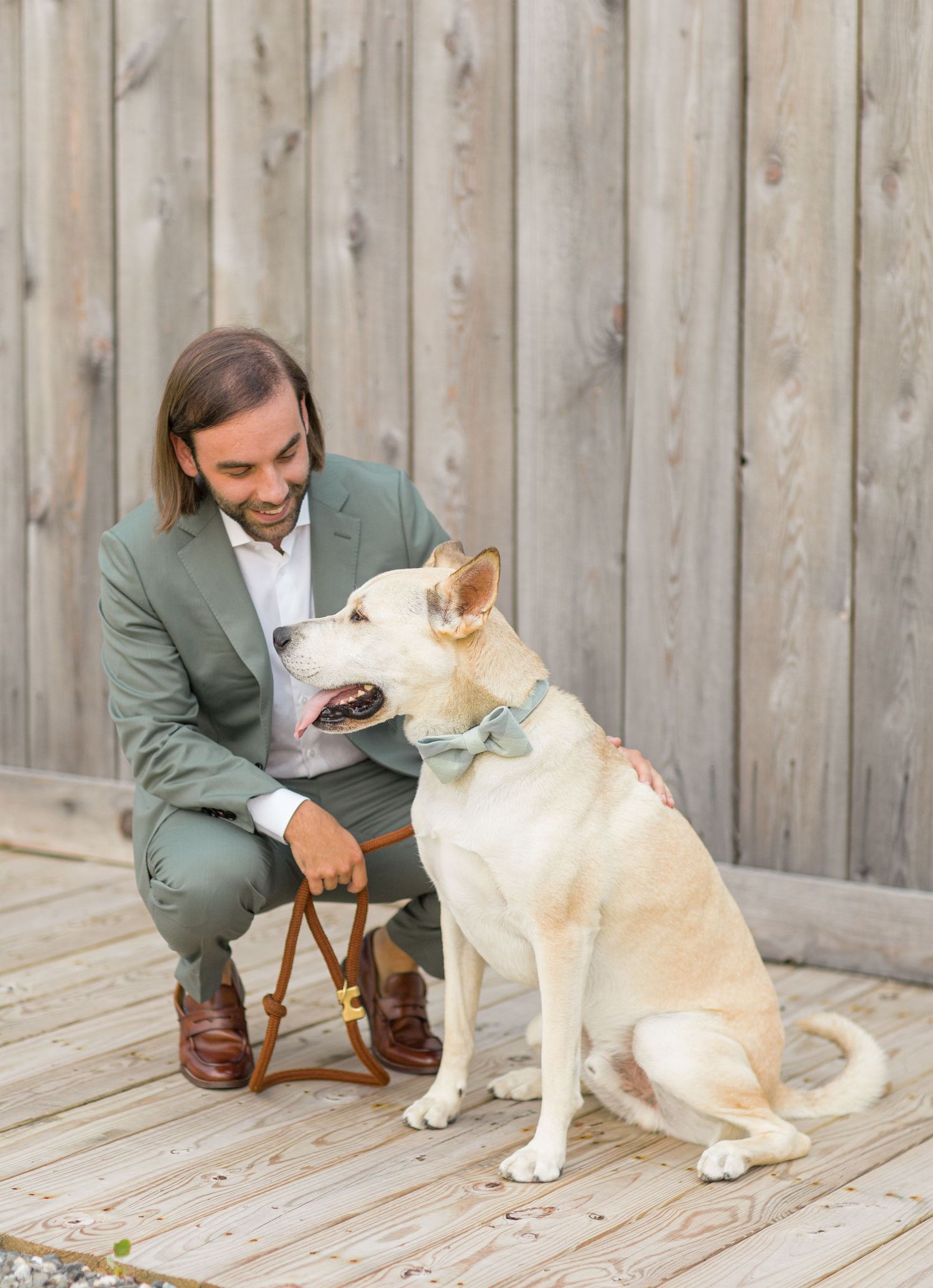Person in green suit jacket sits on wooden deck with a cream-colored dog during a heartfelt interaction.