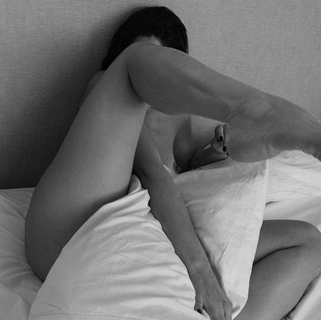 Artistic black and white photograph showing a person reclining on white bedding with dramatic shadowing.