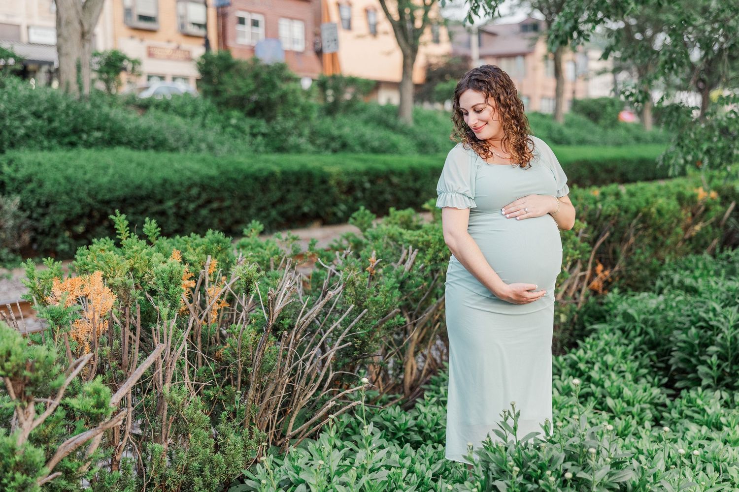Pregnant woman in soft blue dress stands among green foliage during outdoor maternity session.