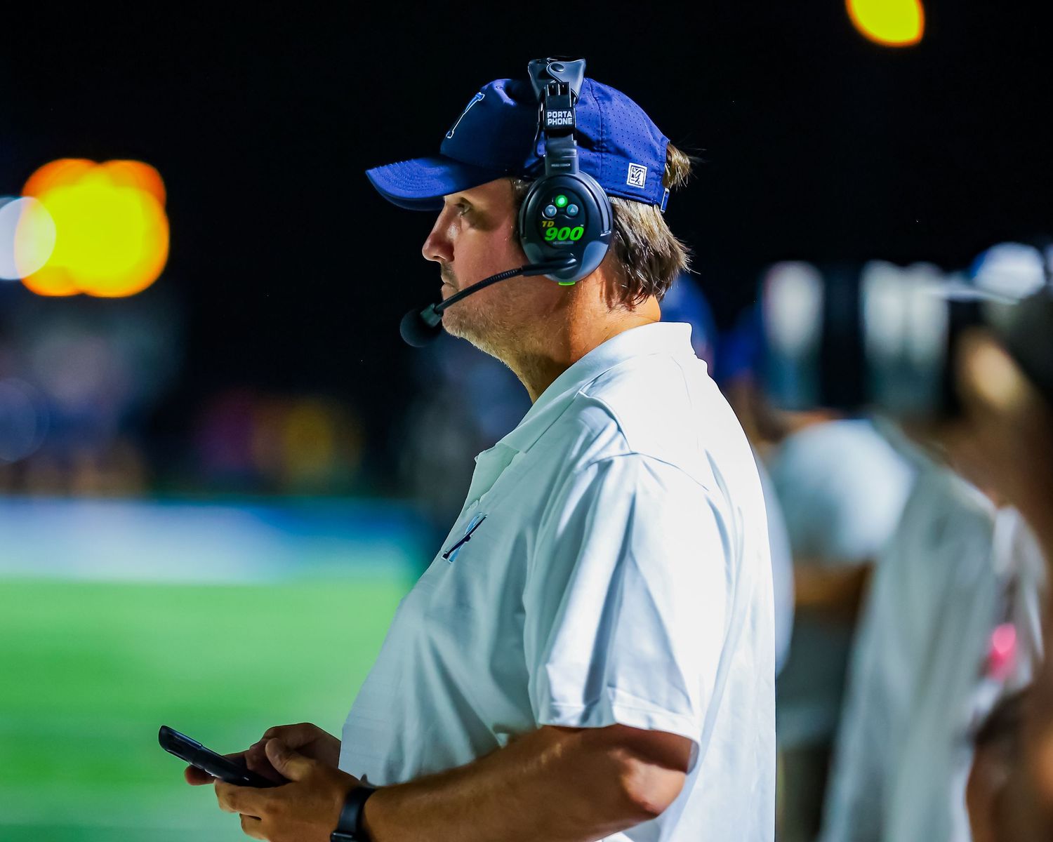 Football coach wearing a blue cap and headset stands on sideline during nighttime game with bokeh lights in background.