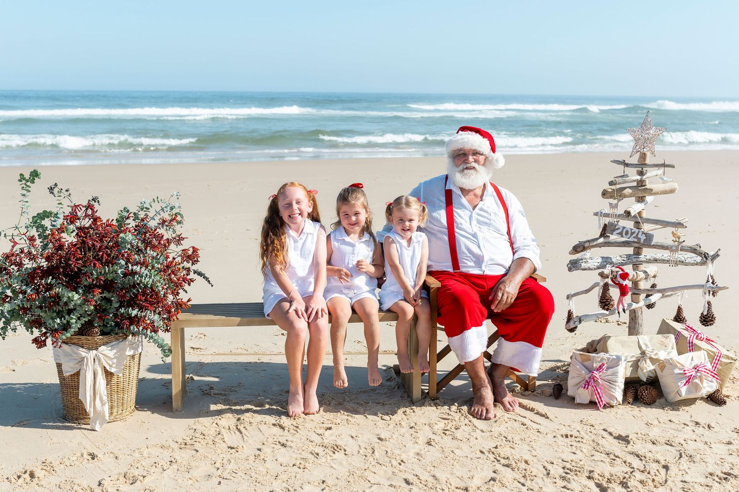 Family members dressed in white sit together on a sandy beach posing with a rustic wooden cross at sunset.
