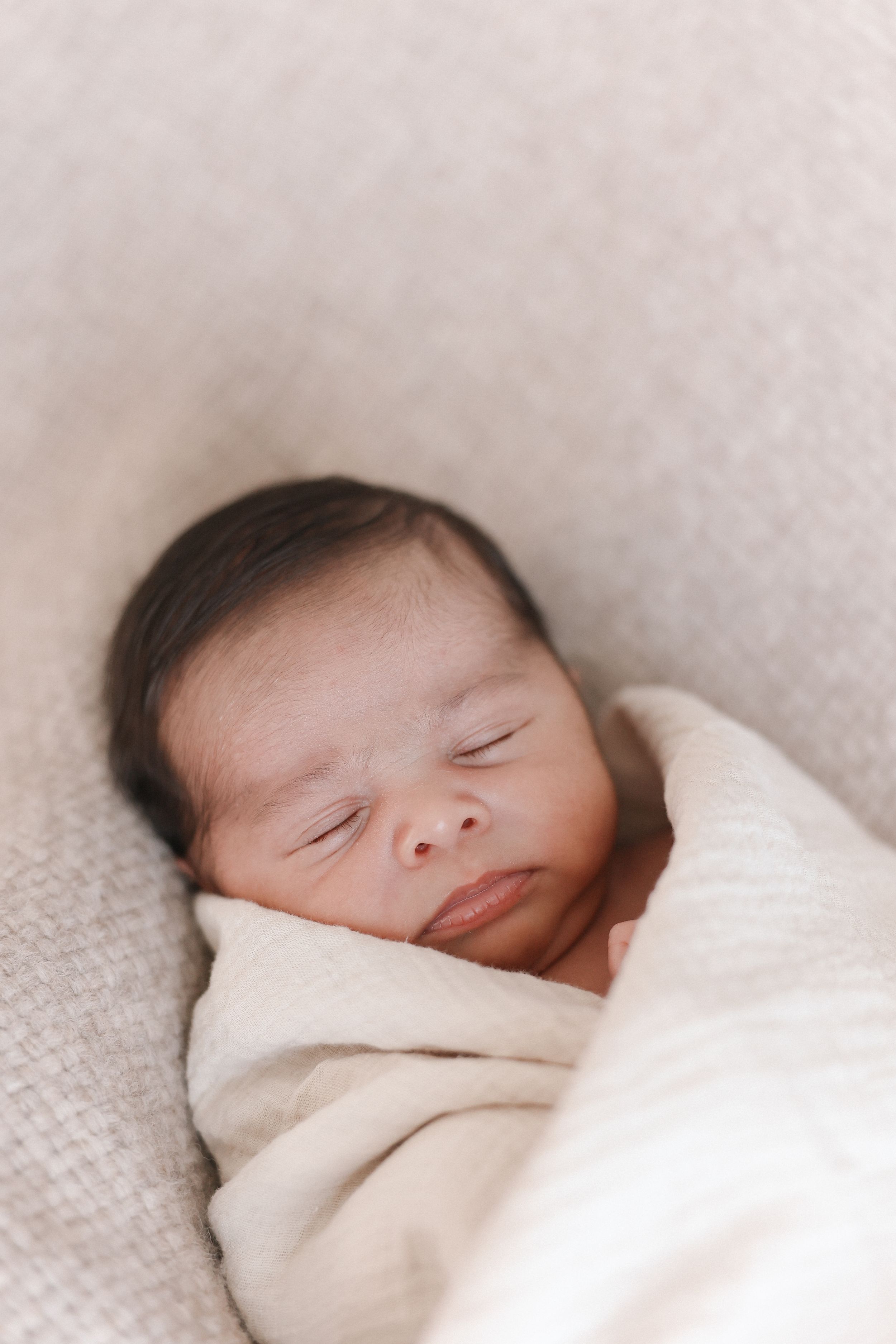 A peaceful newborn baby swaddled in soft white blankets sleeps soundly against a light backdrop.