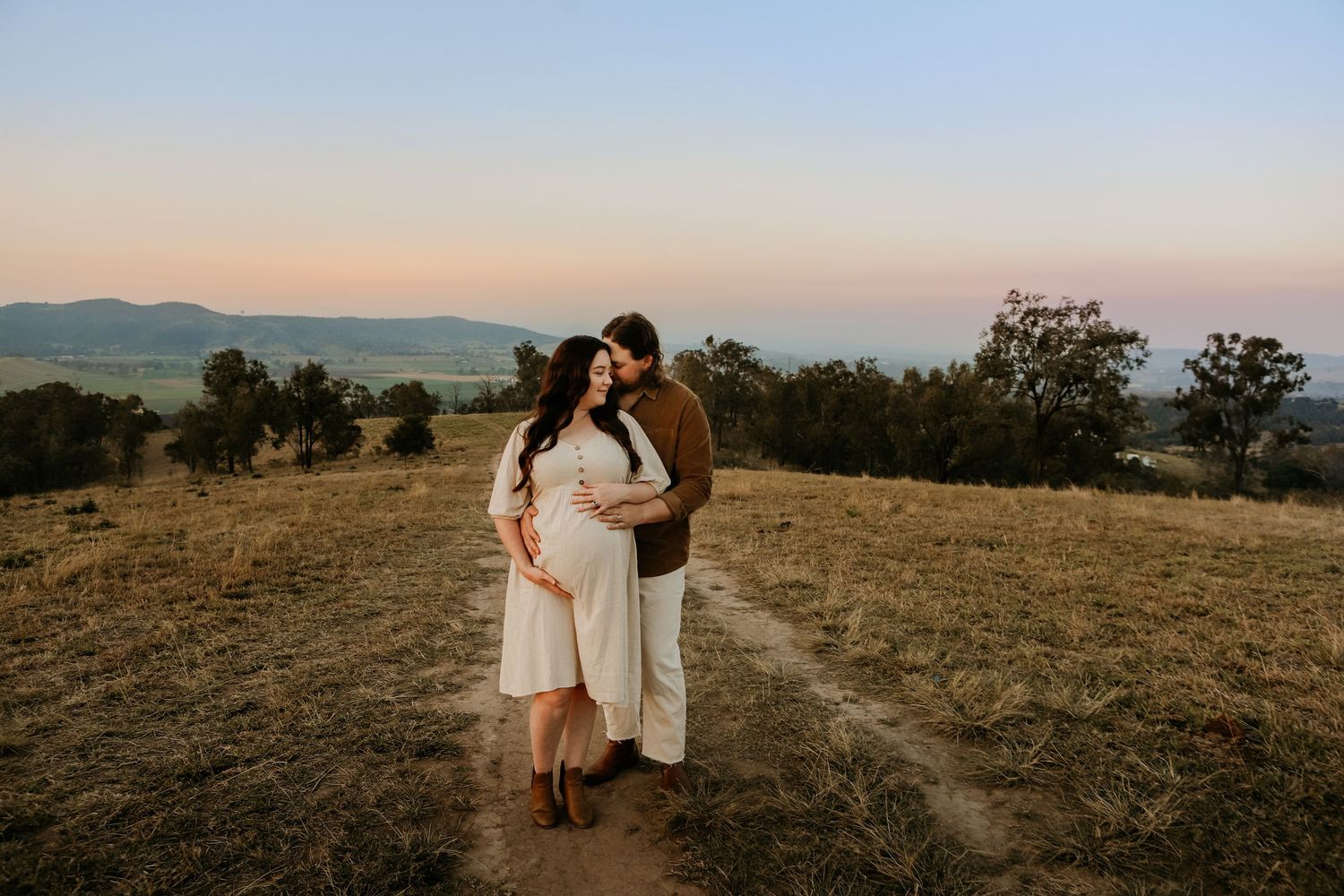 Romantic couple enjoys intimate sunset moment in grassy field with mountain landscape and warm golden light.