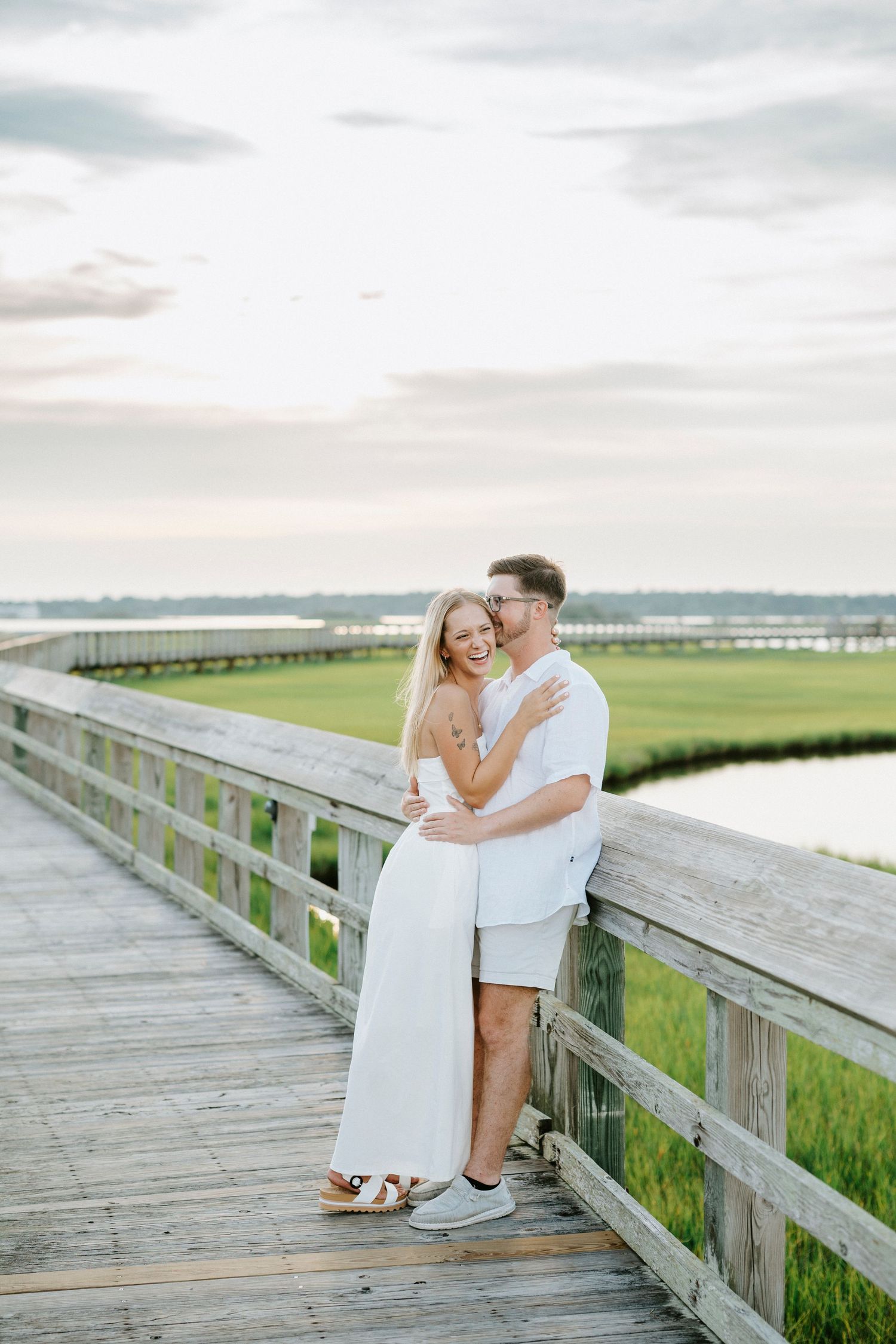 A romantic couple shares tender moments on a wooden boardwalk overlooking a scenic coastal marsh at sunset.