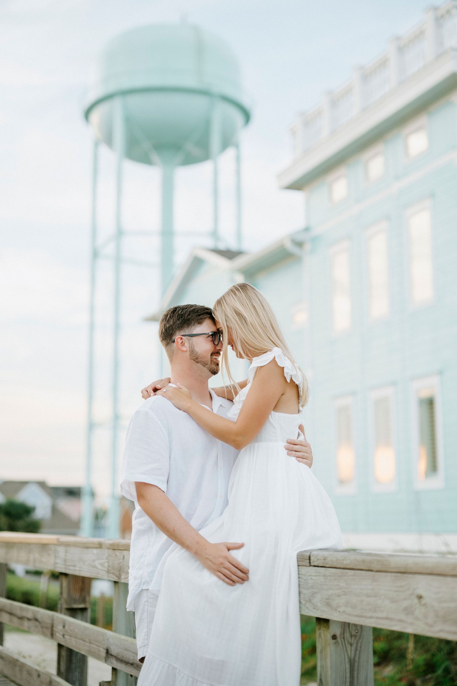 A romantic engagement photo series shows a couple in white outfits embracing at golden hour near a coastal boardwalk.
