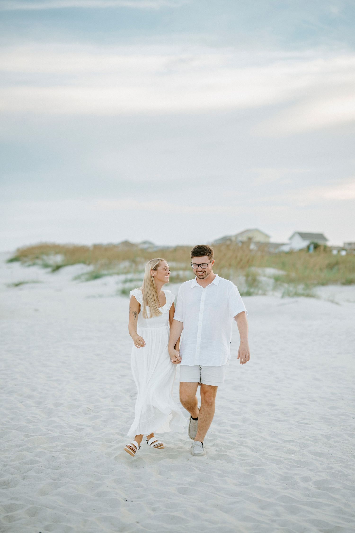 A couple in white outfits walking together along a scenic beach with sand dunes and houses in the background.