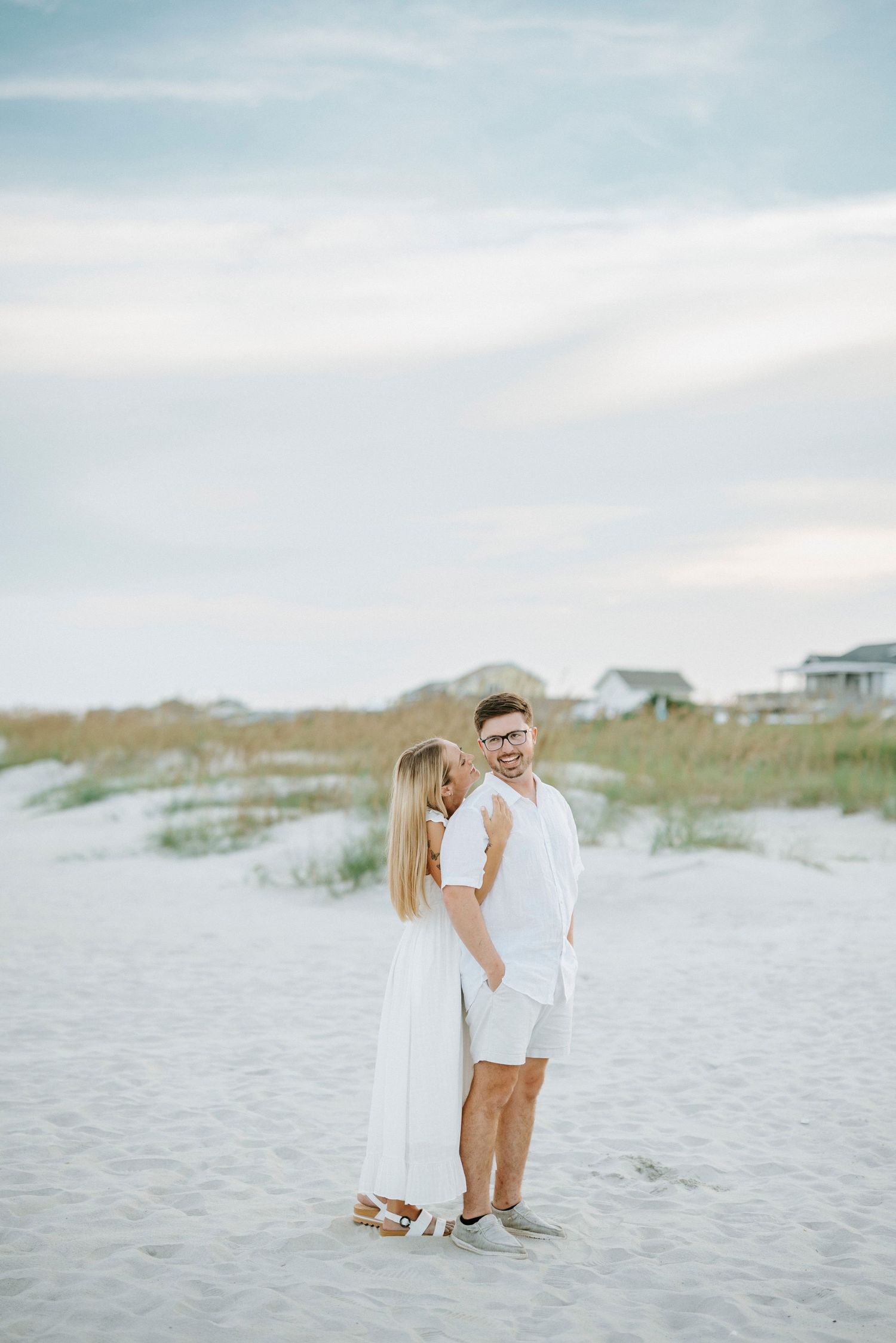 Couple in white outfits embracing on a sandy beach with coastal homes and dunes in the background.