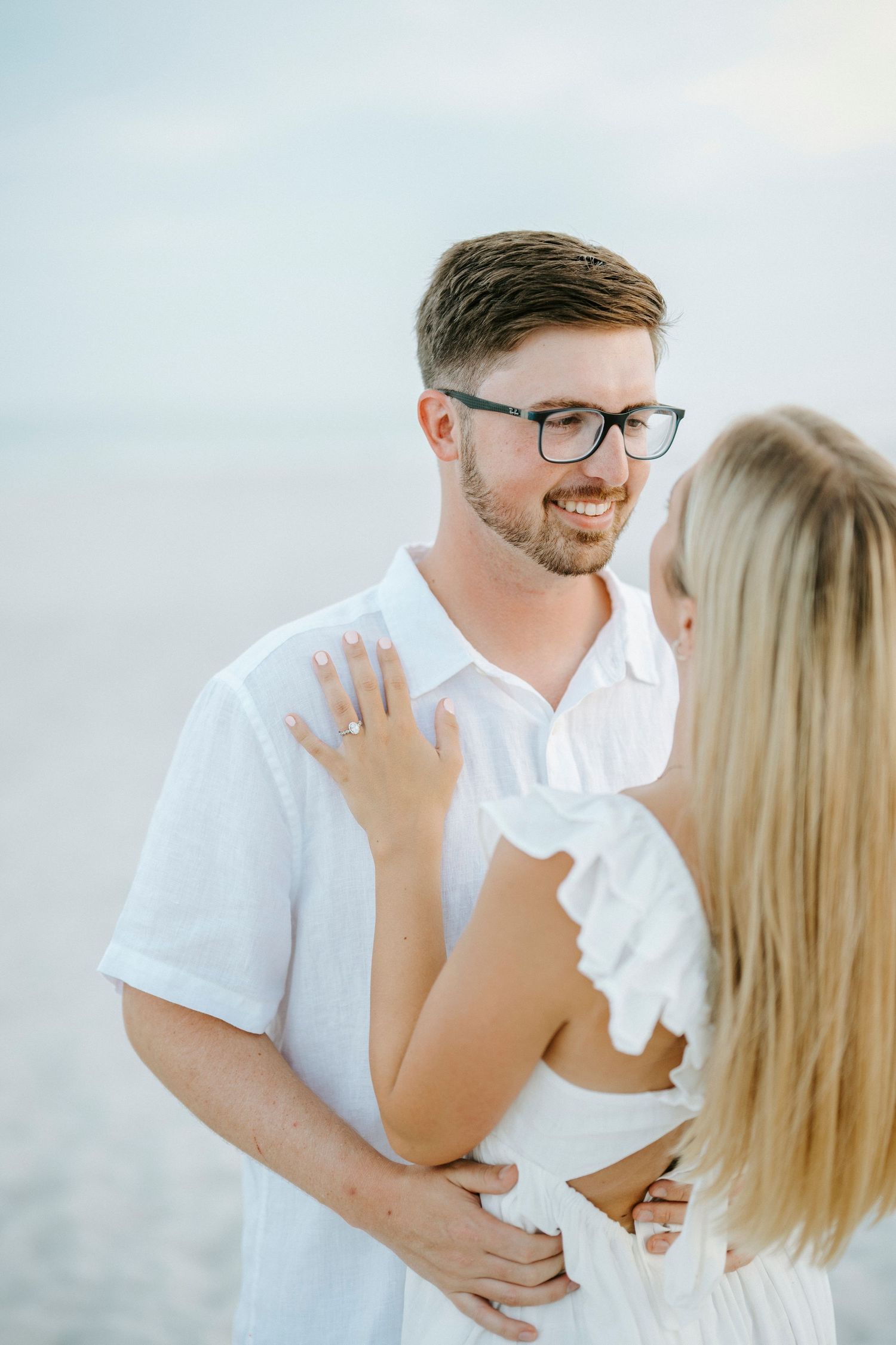A loving couple shares an intimate moment together on a bright, hazy beach wearing white outfits.
