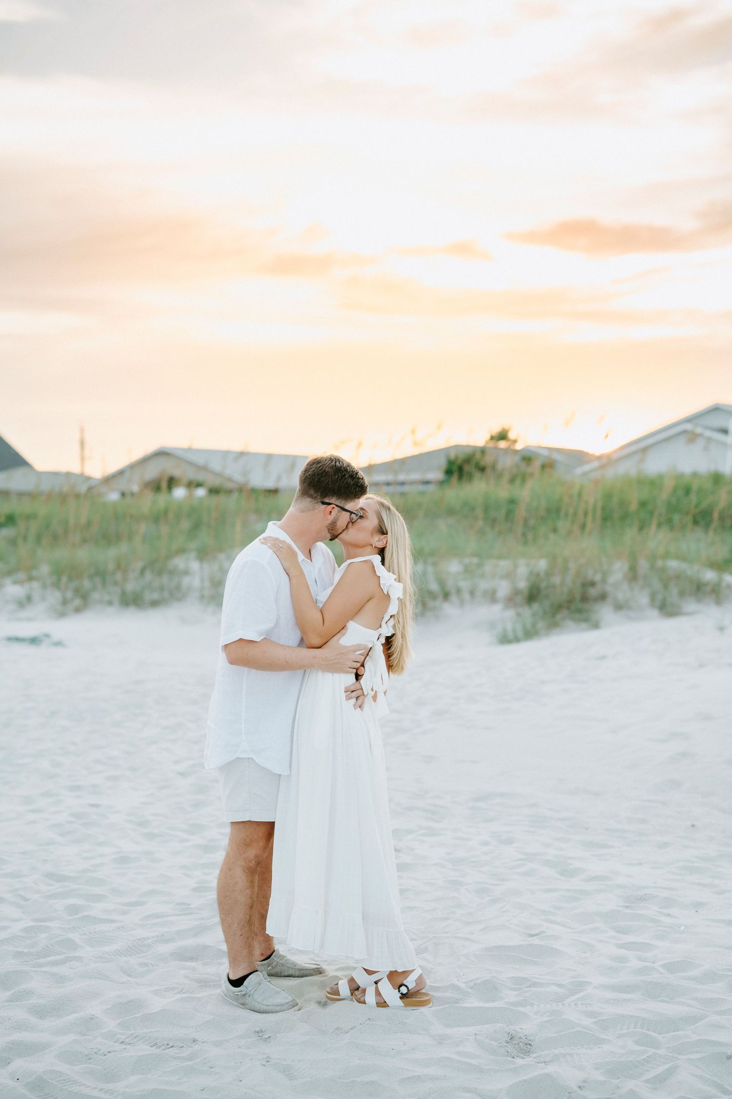 Couple embracing on a sandy beach with sand dunes and warm sunset colors in the background.