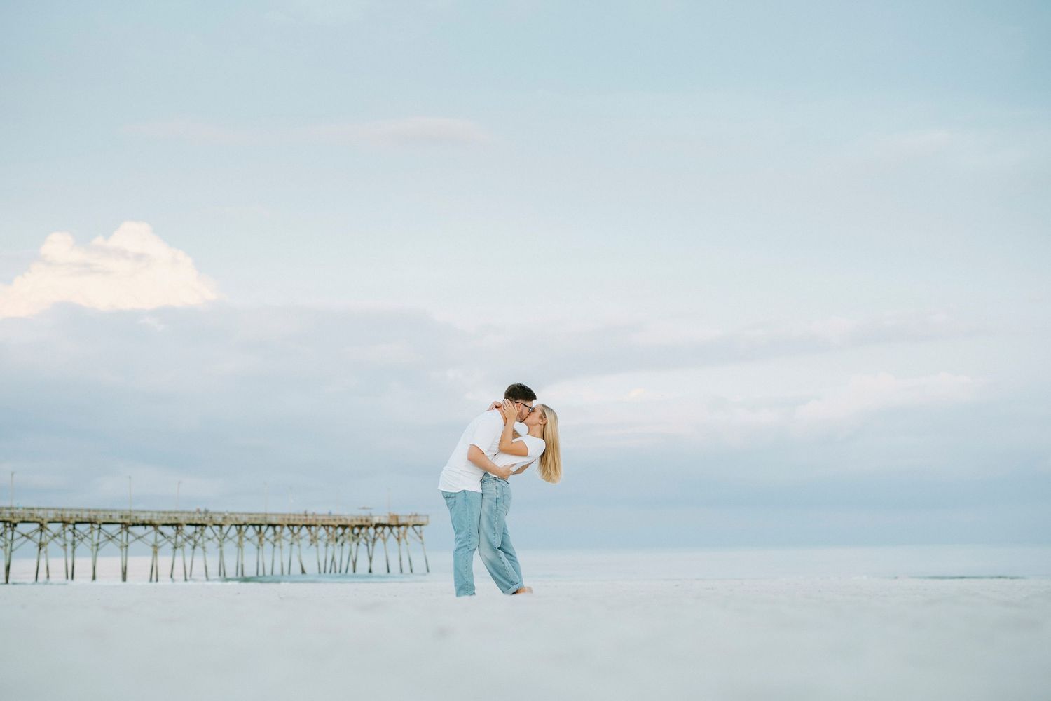 Two figures wearing white embrace on a beach as waves roll in near a wooden pier under a cloudy sky.