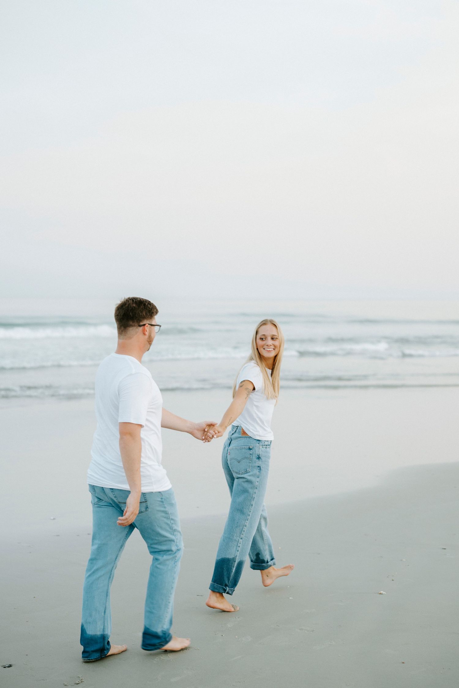 Couple walking barefoot together along a misty beach shoreline in casual denim and white attire.