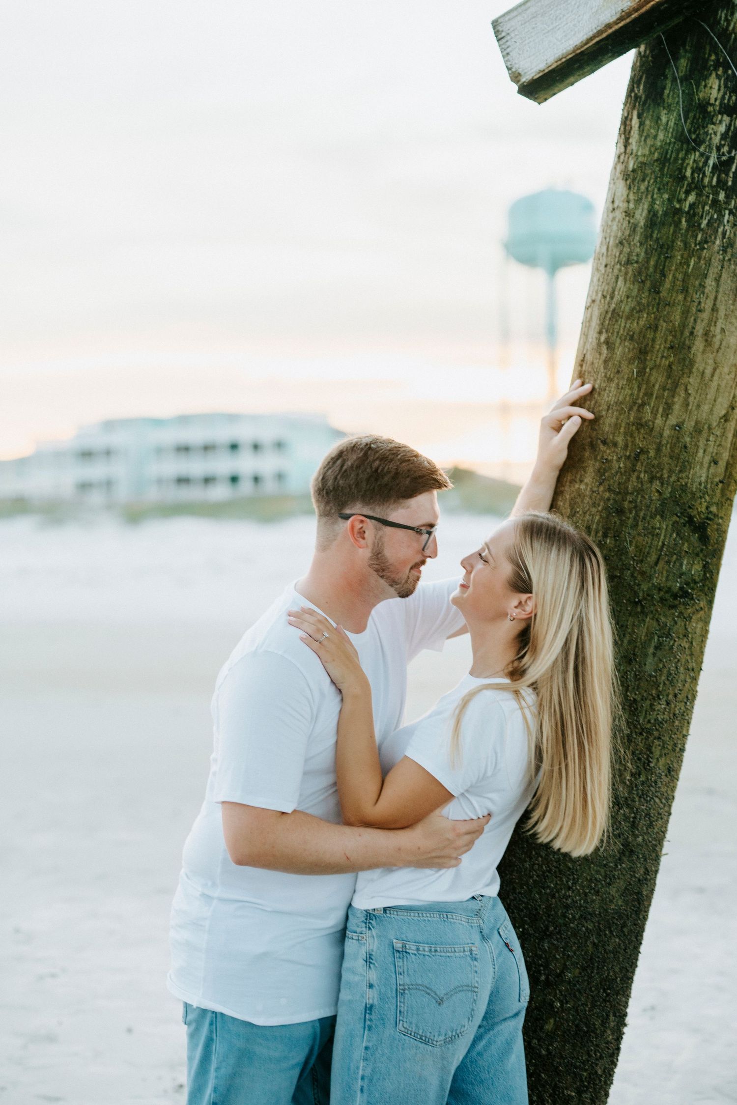 Romantic couple wearing white and denim embrace by wooden posts on a sunny beach.