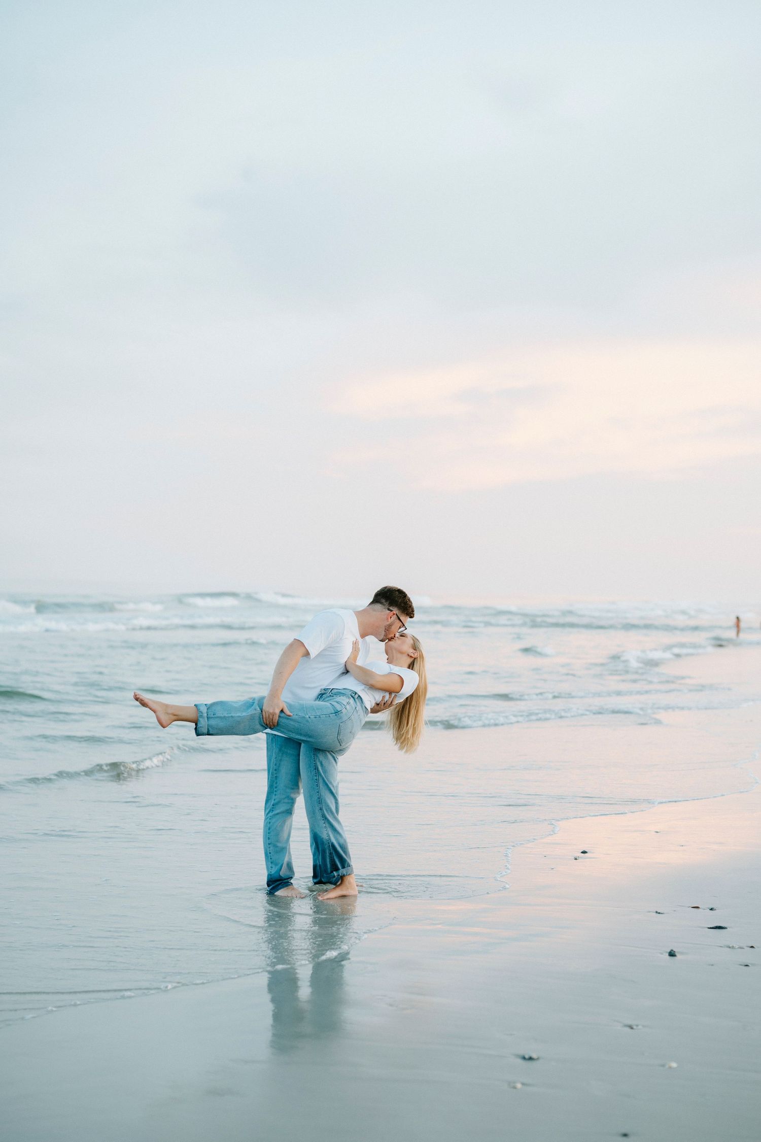 A couple dips and poses playfully in the ocean waves at sunset, creating a romantic beach silhouette.