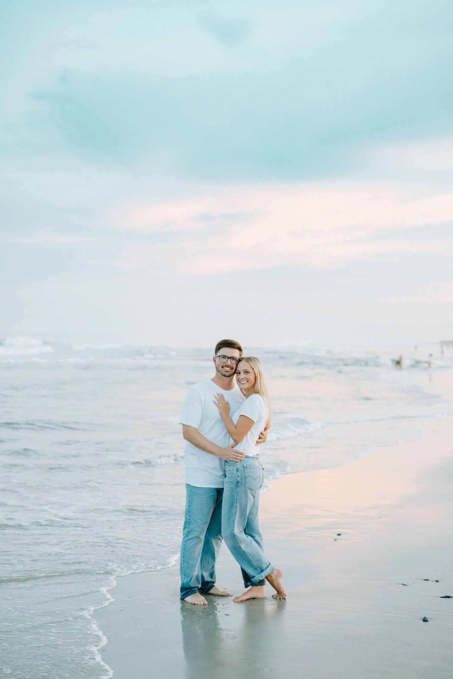 A couple embraces on the beach during sunset wearing white and blue casual attire along the ocean shoreline.
