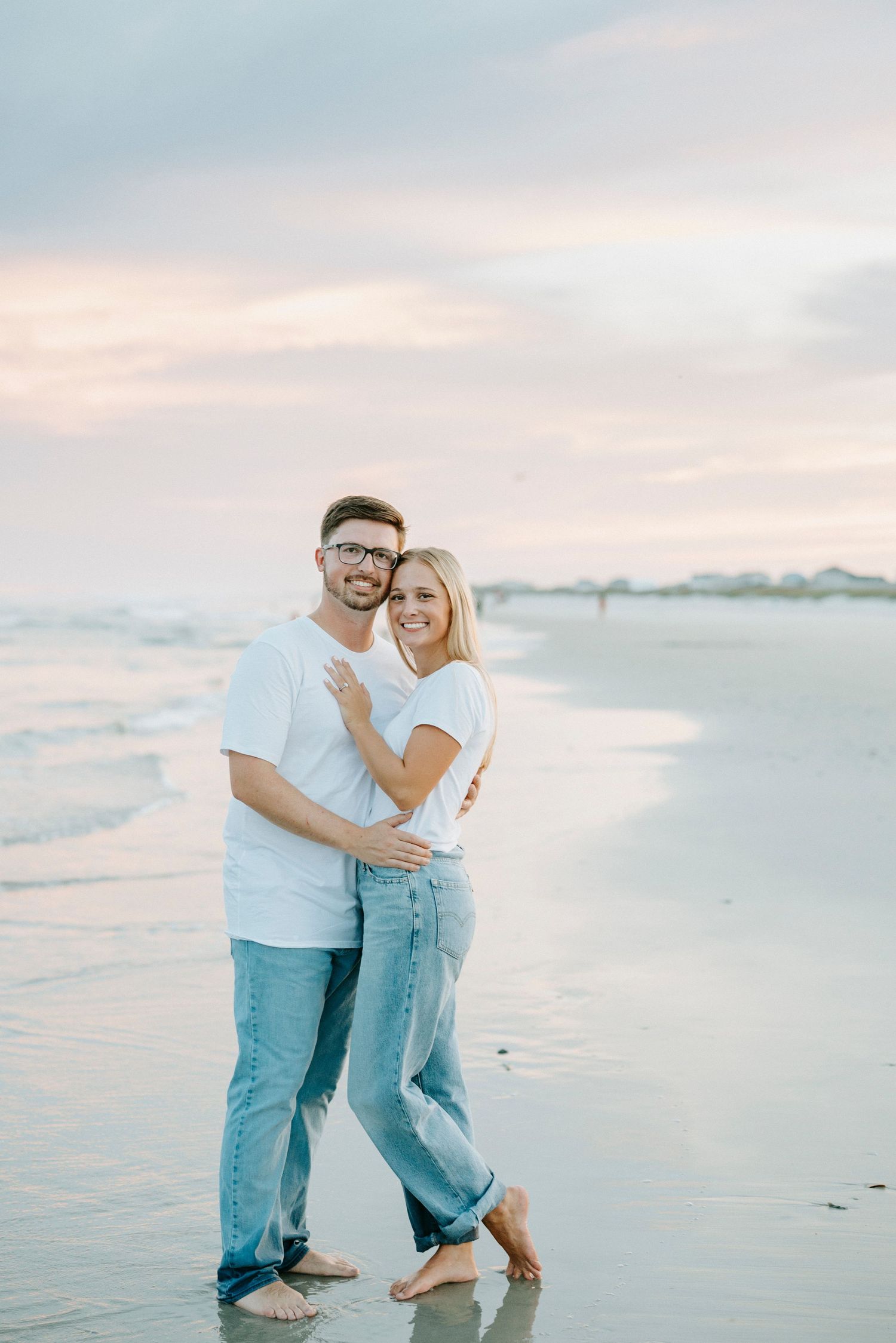 Two people embrace on a serene beach at sunset with calm ocean waters and warm lighting.
