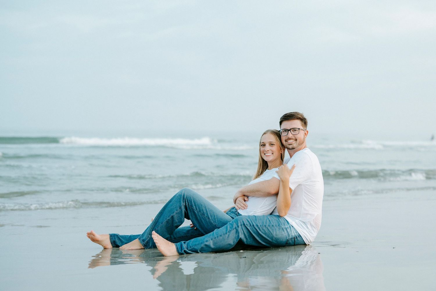 Romantic couple poses on sandy beach with waves in background, creating dreamy coastal engagement photos.