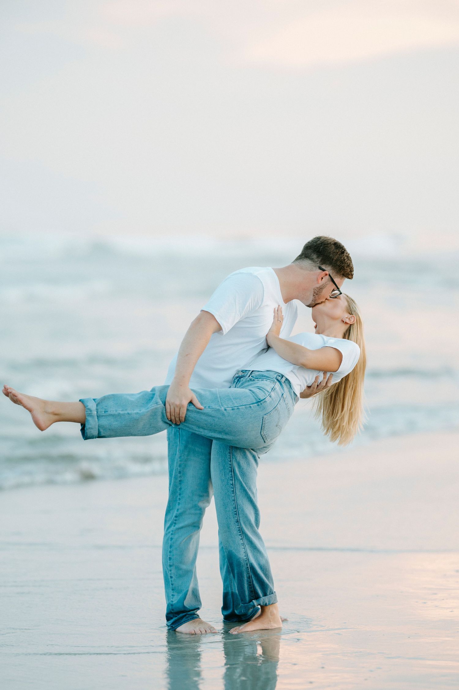 A couple shares a romantic dance-like pose in the shallow ocean waters during a dreamy sunset photoshoot.