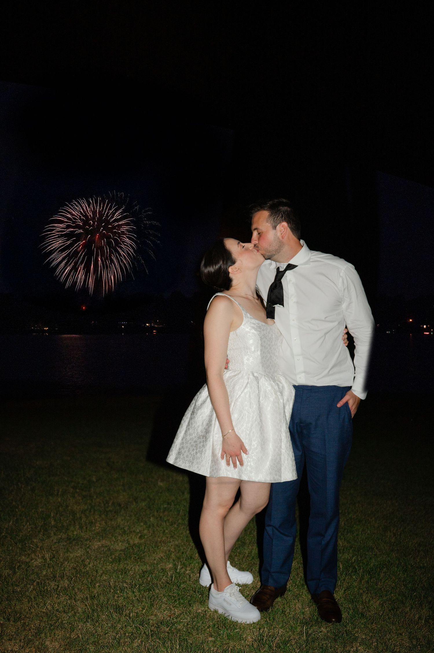 A newlywed couple shares a romantic kiss at night while fireworks burst in the dark sky behind them. Photo by Joliette Marie Photography.