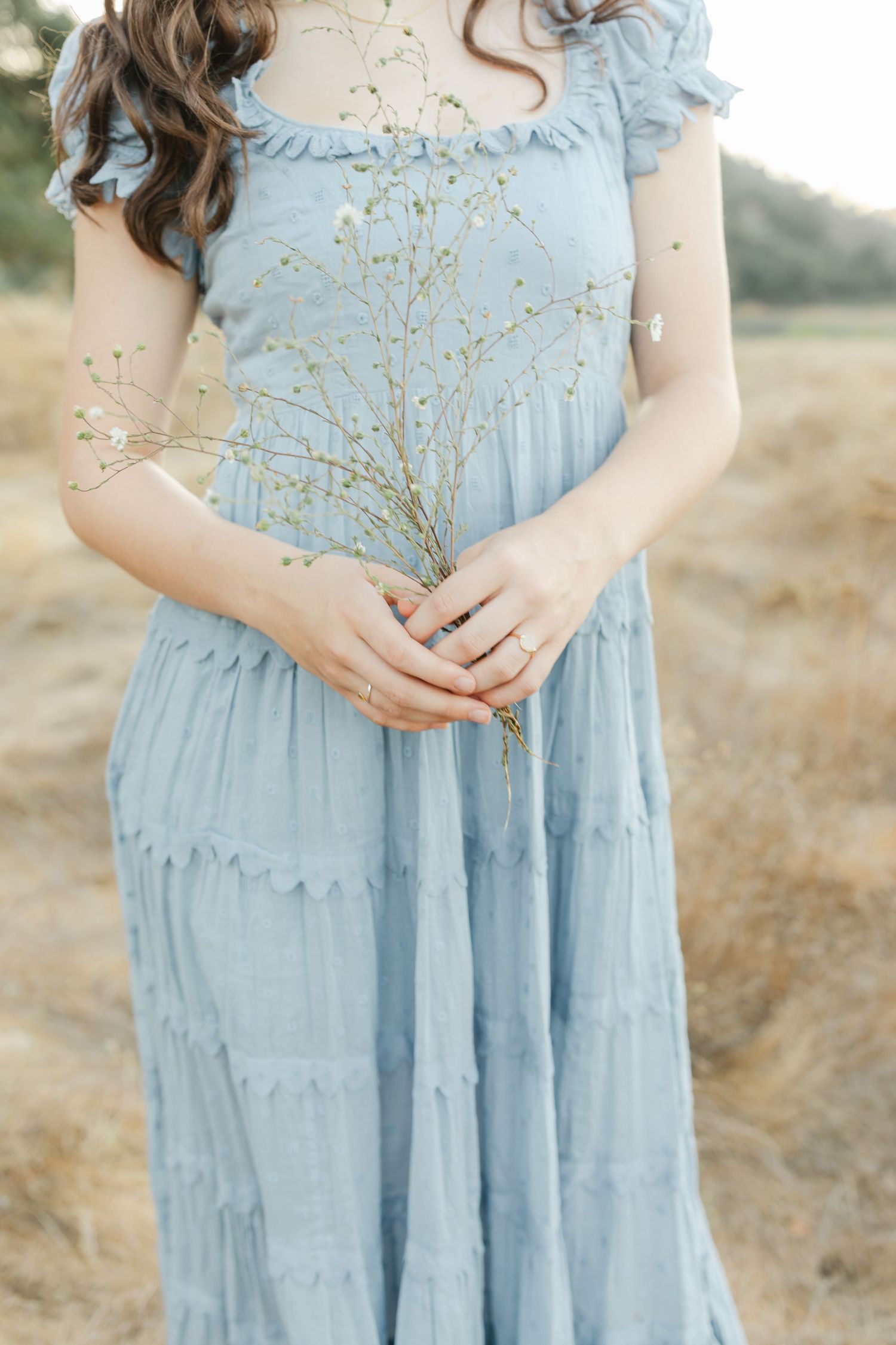 A person in a light blue vintage-style dress with ruffled details poses outdoors in a golden field.Film by Chelsee Rawe Photography