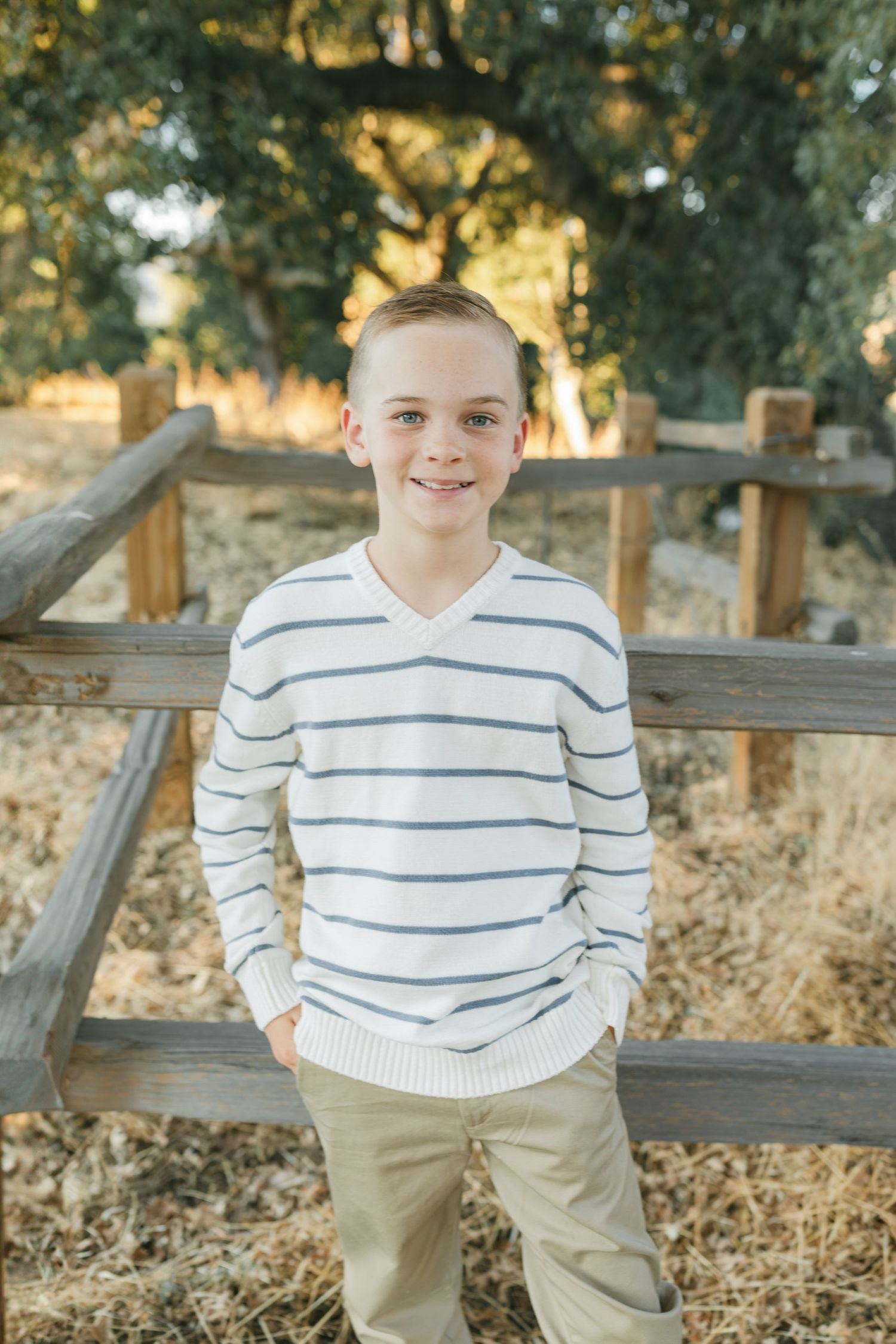 Young child in striped white and blue sweater and khaki pants sits on rustic wooden fence in autumn setting captured by Chelsee Rawe Photog.