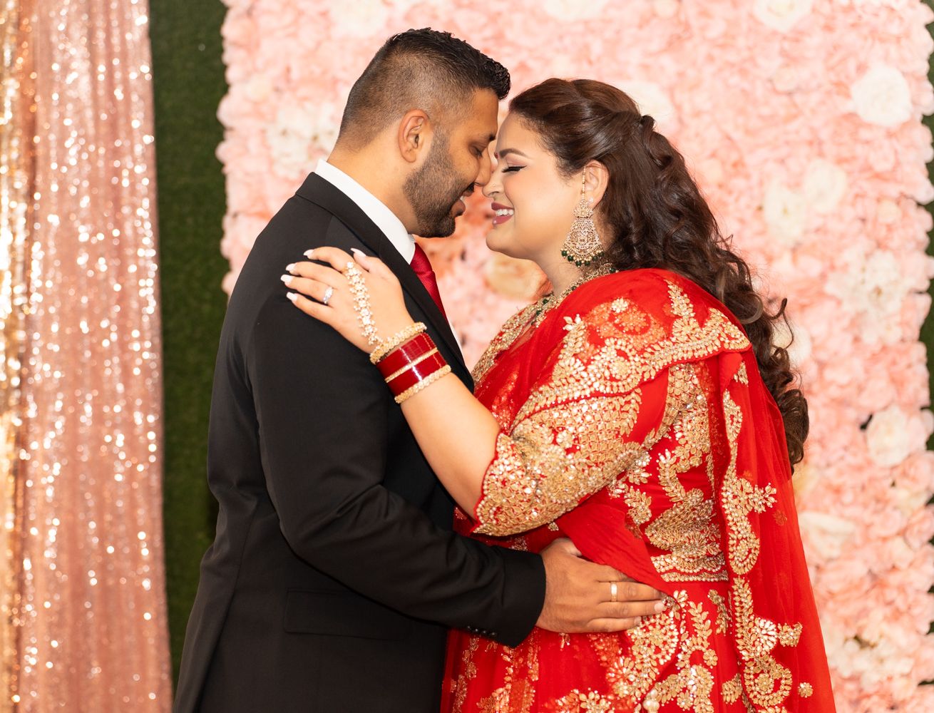 Indian wedding celebration with couple in traditional red attire embracing against a champagne sequin backdrop.