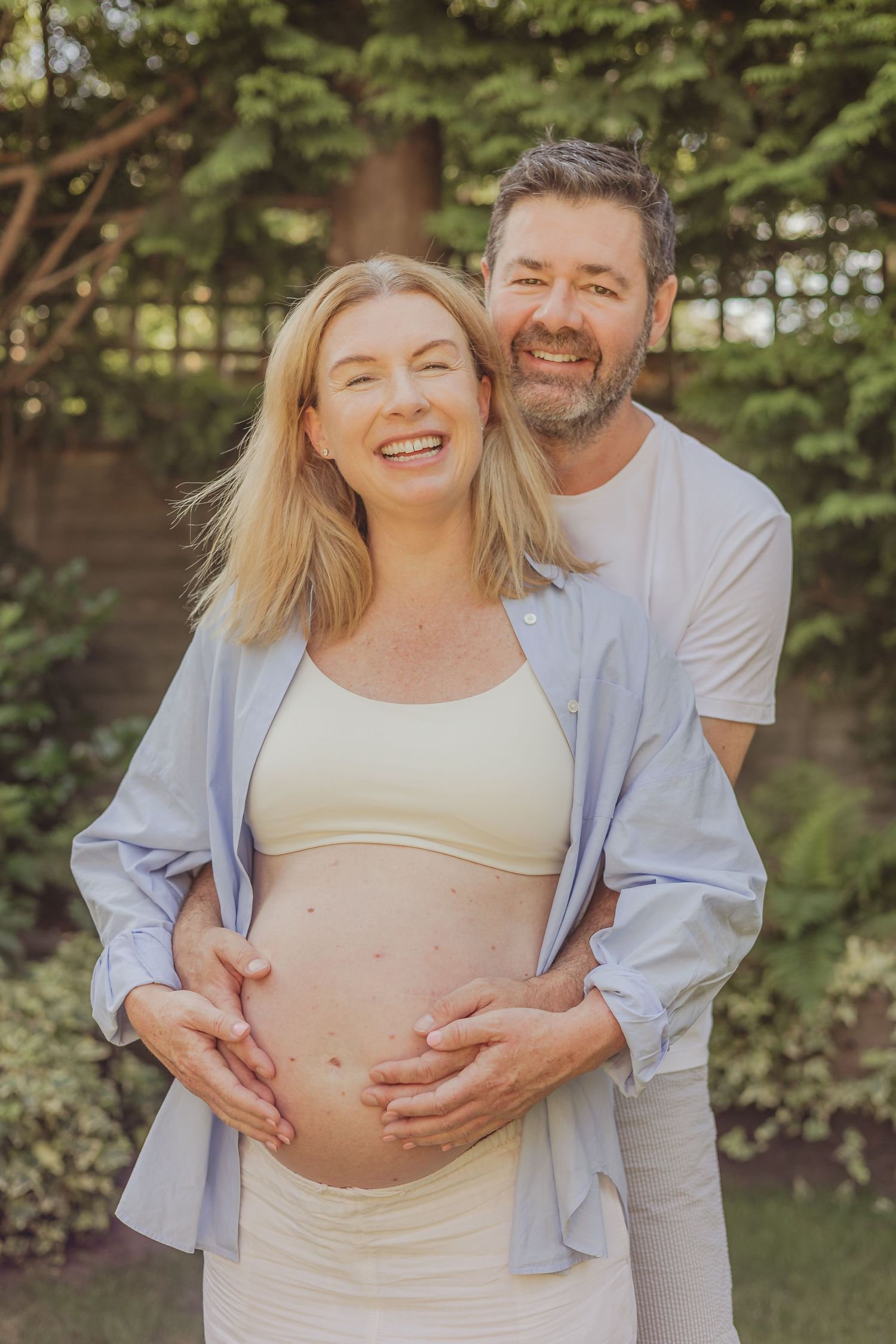 Intimate outdoor maternity photo with couple embracing in front of green foliage.