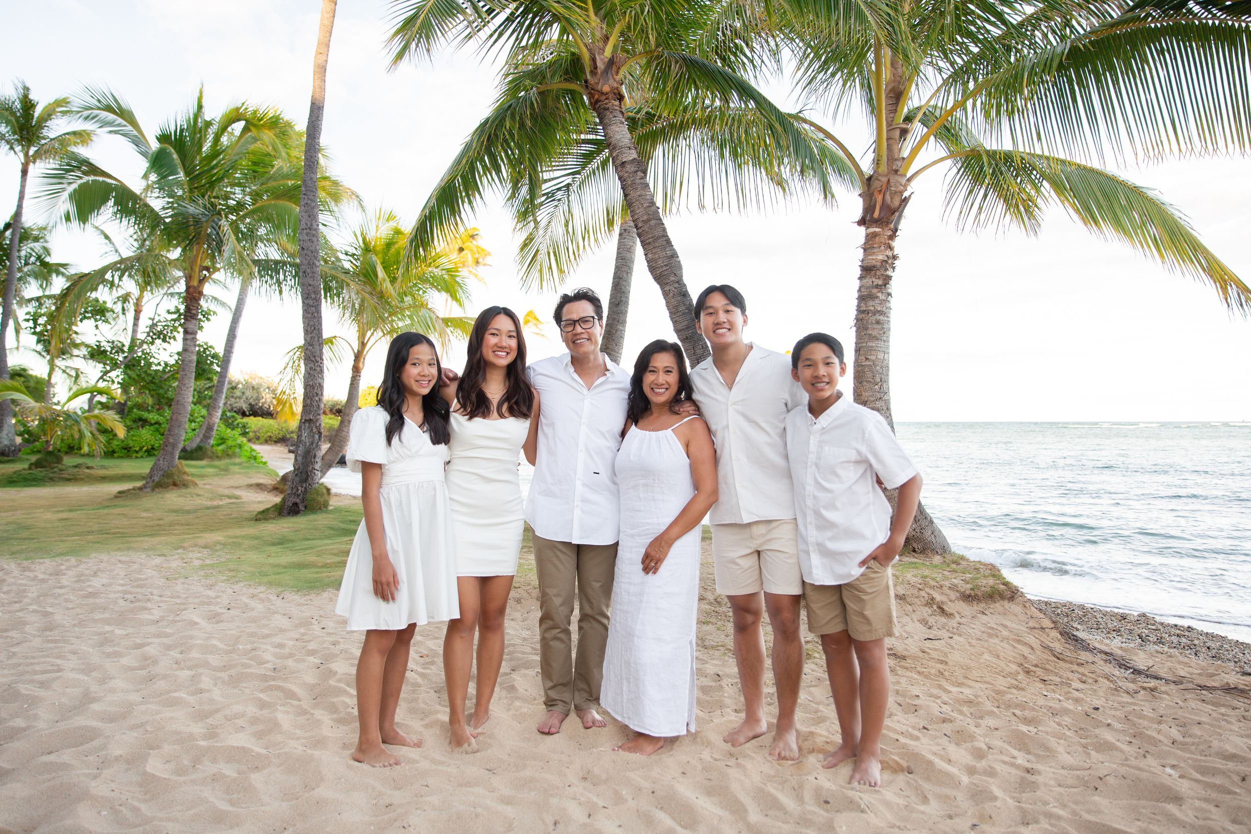 A group in white outfits posing for a series of family portraits on a tropical beach with palm trees and ocean views at Kahala Hotel.
