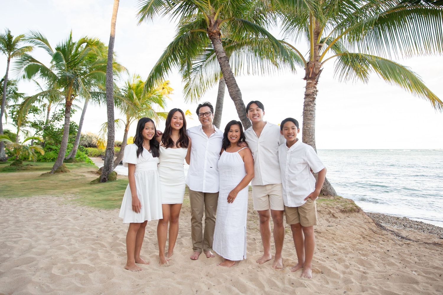 A group in white outfits posing for a series of family portraits on a tropical beach with palm trees and ocean views at Kahala Hotel.