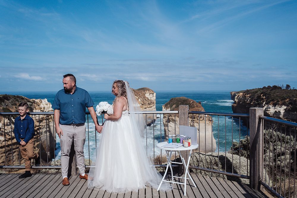 Couple in wedding attire walks along scenic oceanside boardwalk on bright sunny day with cliffs in background.