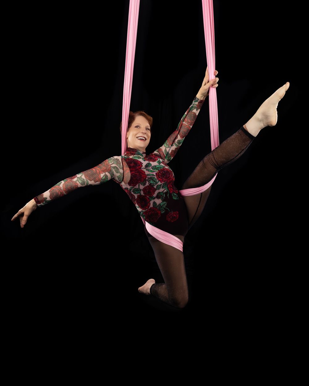 An aerial performer executes a dynamic routine on pink silk ribbons against a dark background.