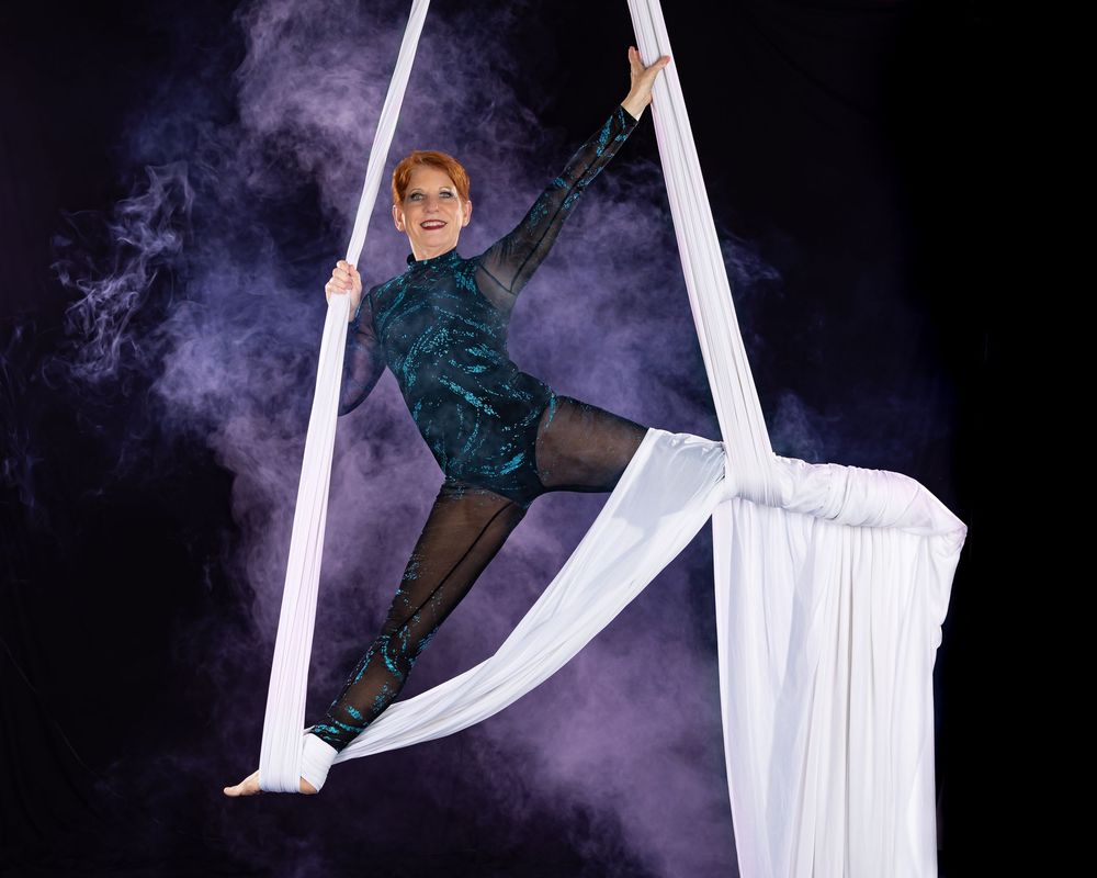 Aerial silk performer in dark costume gracefully poses on white fabric against dramatic purple smoke background.
