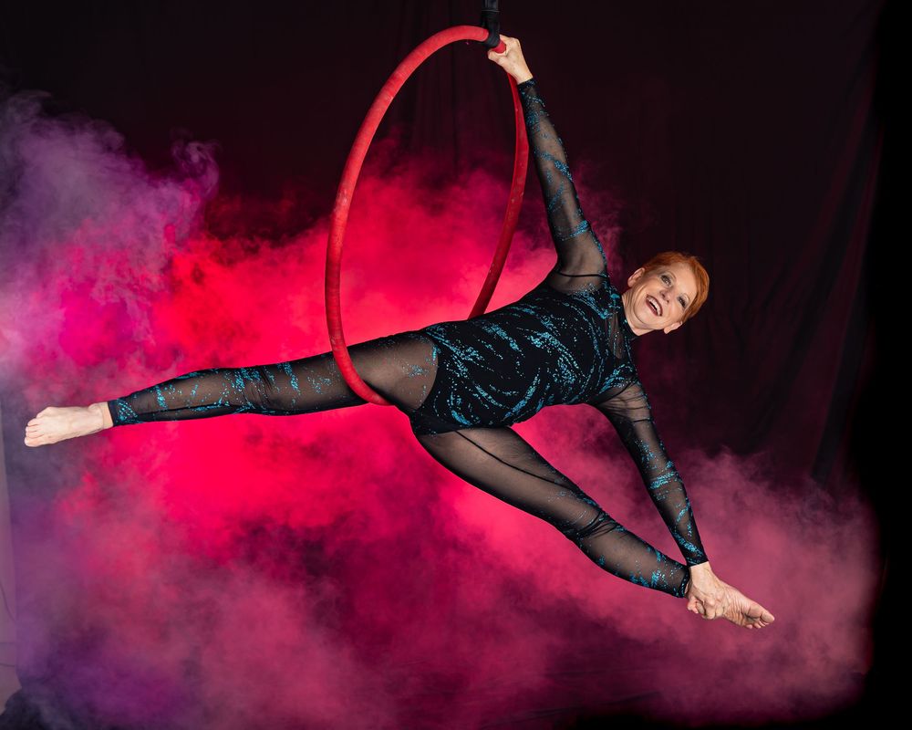 Aerial dancer performing suspended movements in black outfit against vibrant red and purple smoky backdrop.