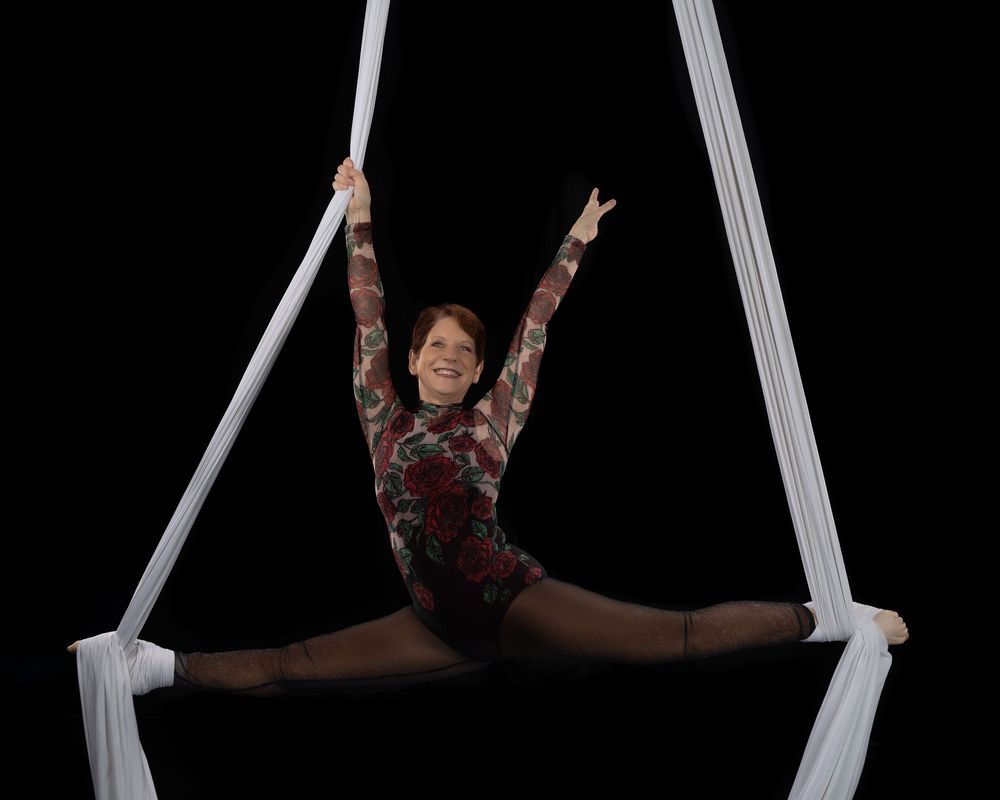 Aerial dancer performs a split while suspended from white silks against a black background, showing graceful movement.