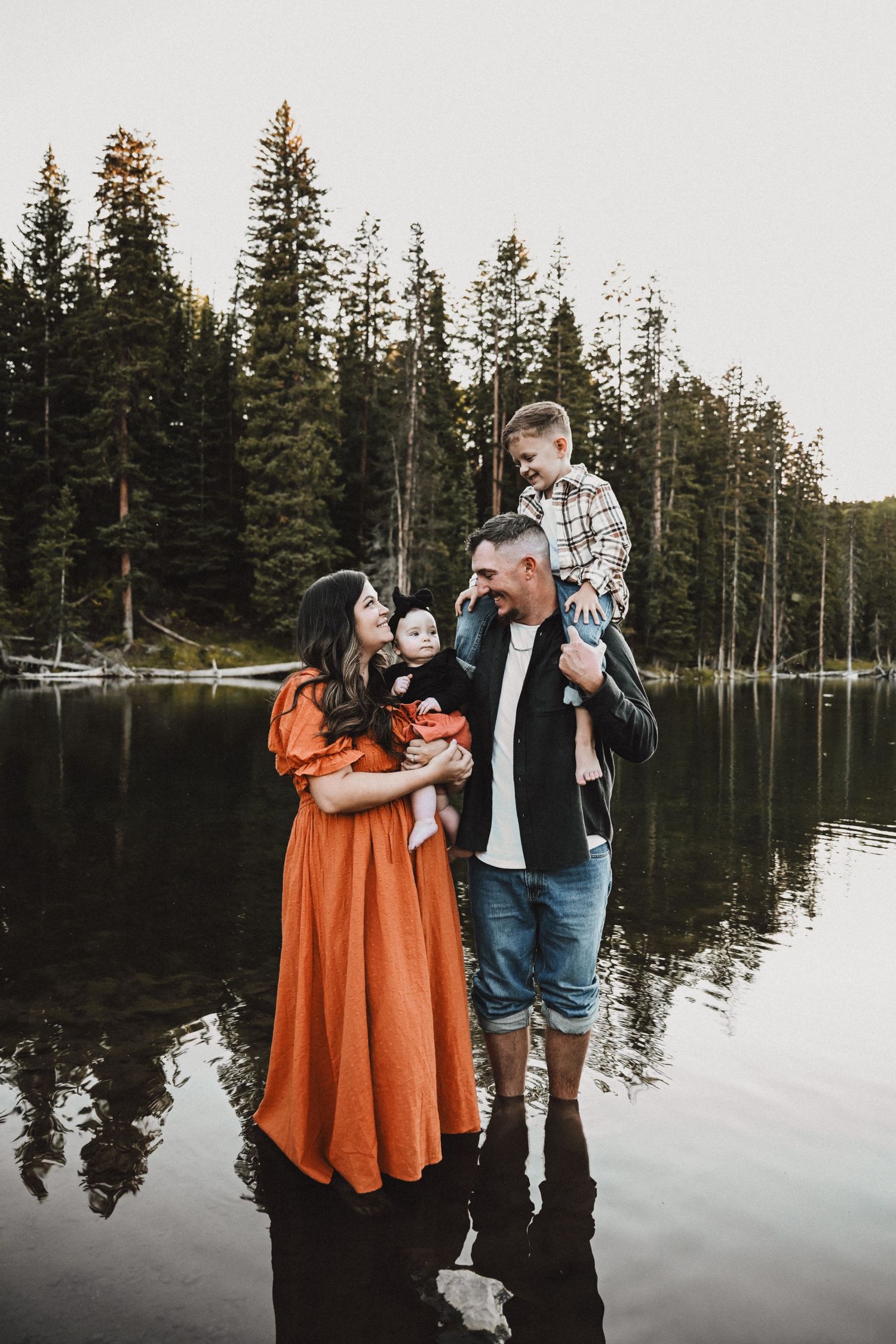 Couple poses playfully by a lake with evergreen trees, wearing orange and denim outfits.