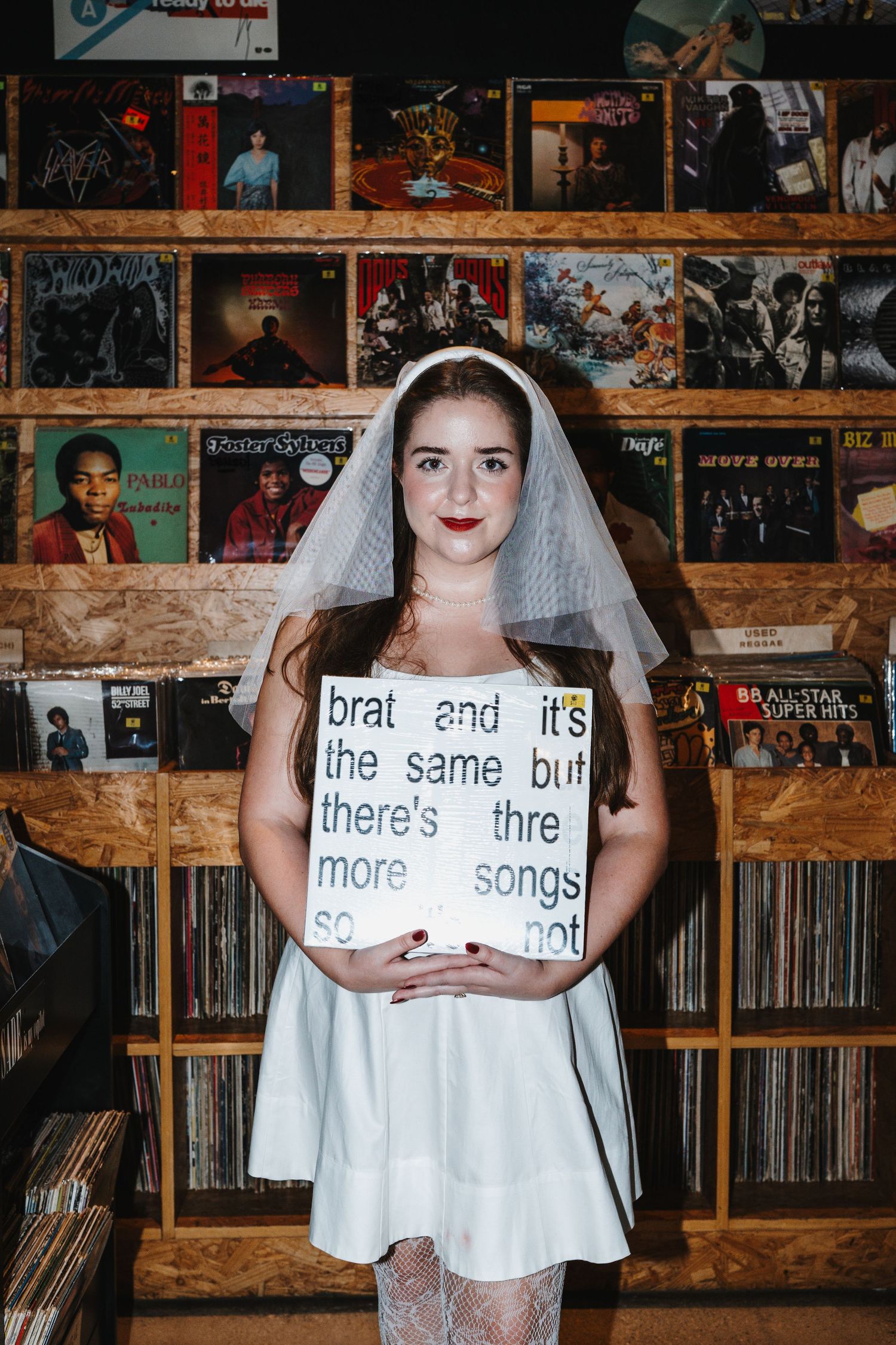 Someone in a bridal veil holds up signs with text in a room filled with vintage records and album covers on brick walls.