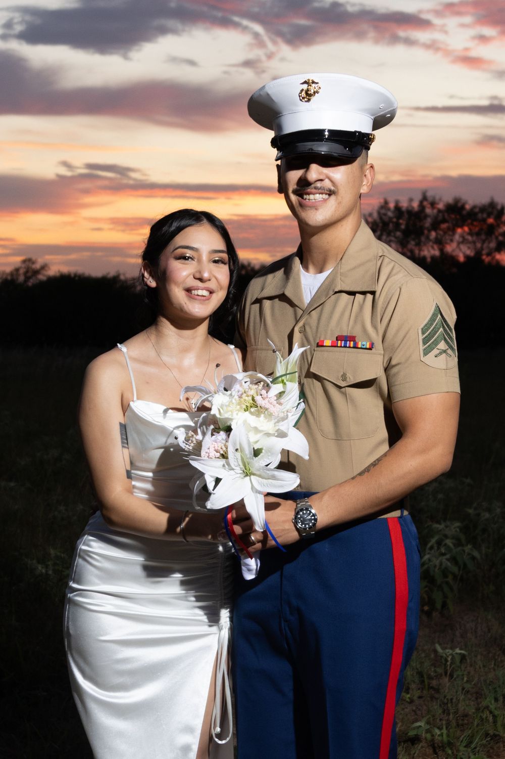 A wedding couple poses at sunset, with the groom in Marine dress uniform and bride holding a bouquet of white lilies.