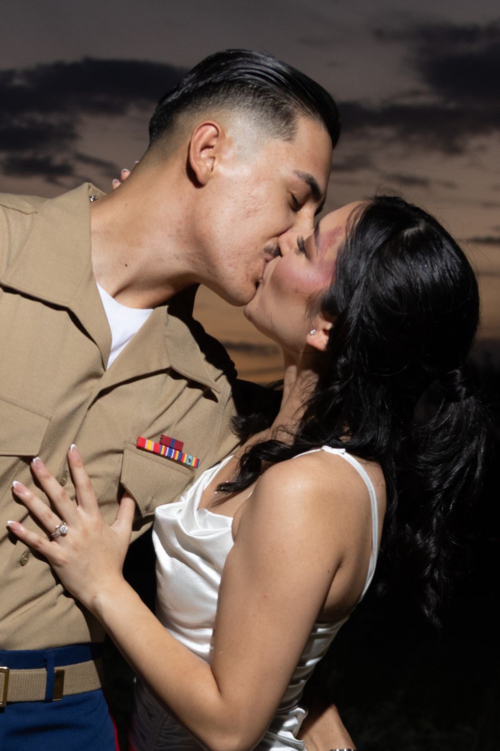 A couple shares a passionate kiss in front of a dramatic sunset sky while he wears military attire and she wears white.