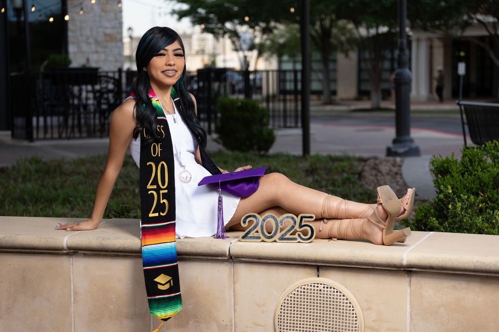 A graduate in festive graduation regalia with decorative sash and stole sits casually on a stone wall outdoors.