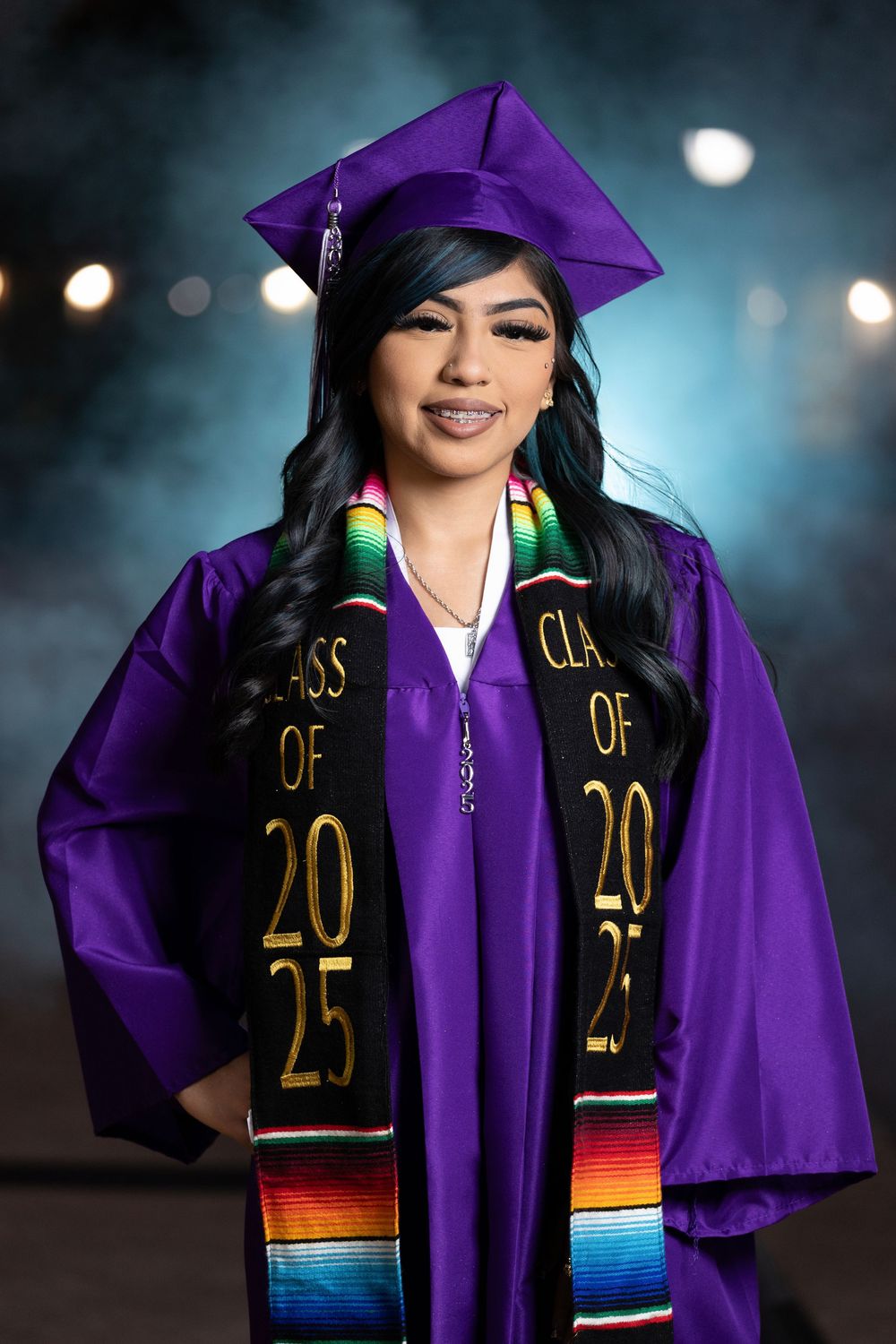Graduate wearing purple cap and gown with colorful Class of 2023 stole poses for graduation photos at dusk.