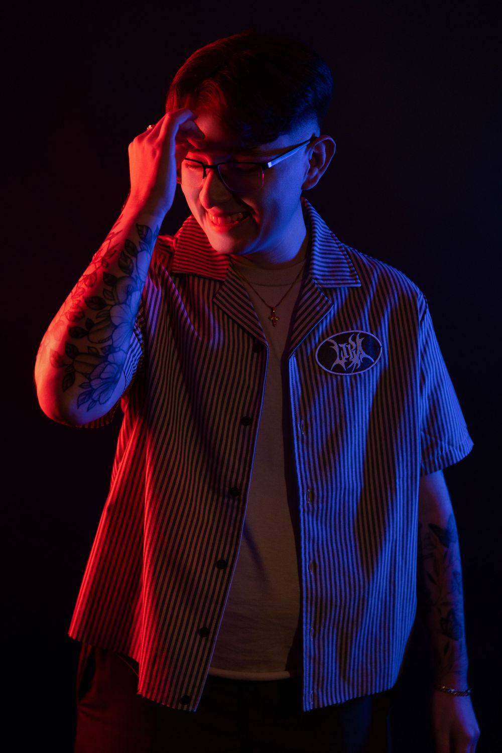 Figure lit dramatically with red and blue lighting wearing striped jacket and white shirt in dark studio setting.