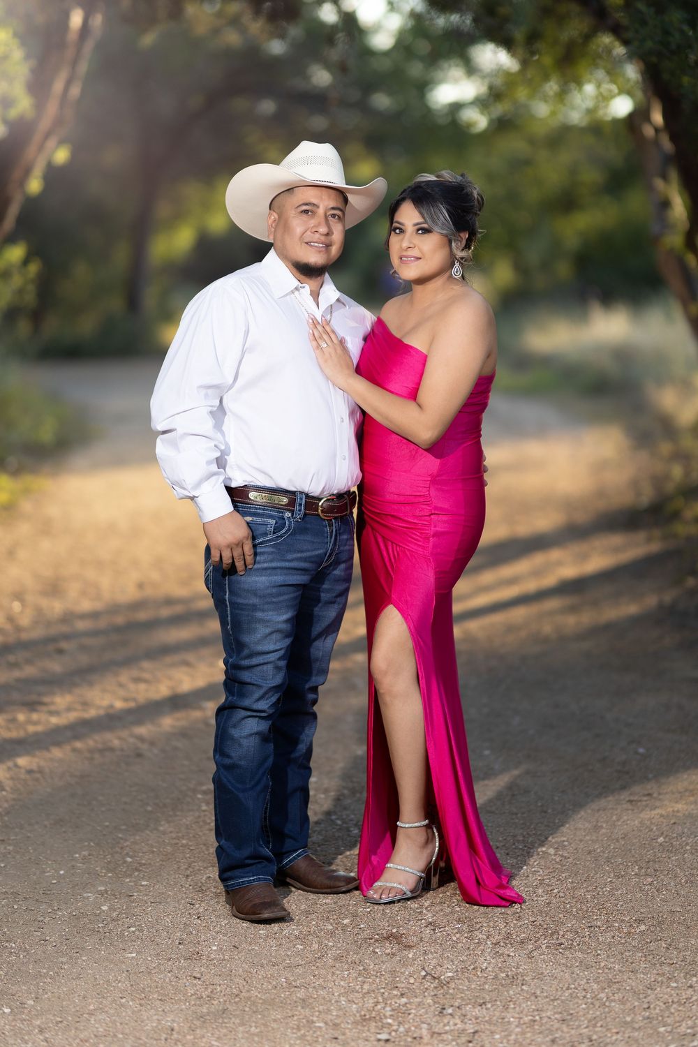A couple poses together outdoors, with one person wearing white and jeans and the other in a vibrant pink formal dress.